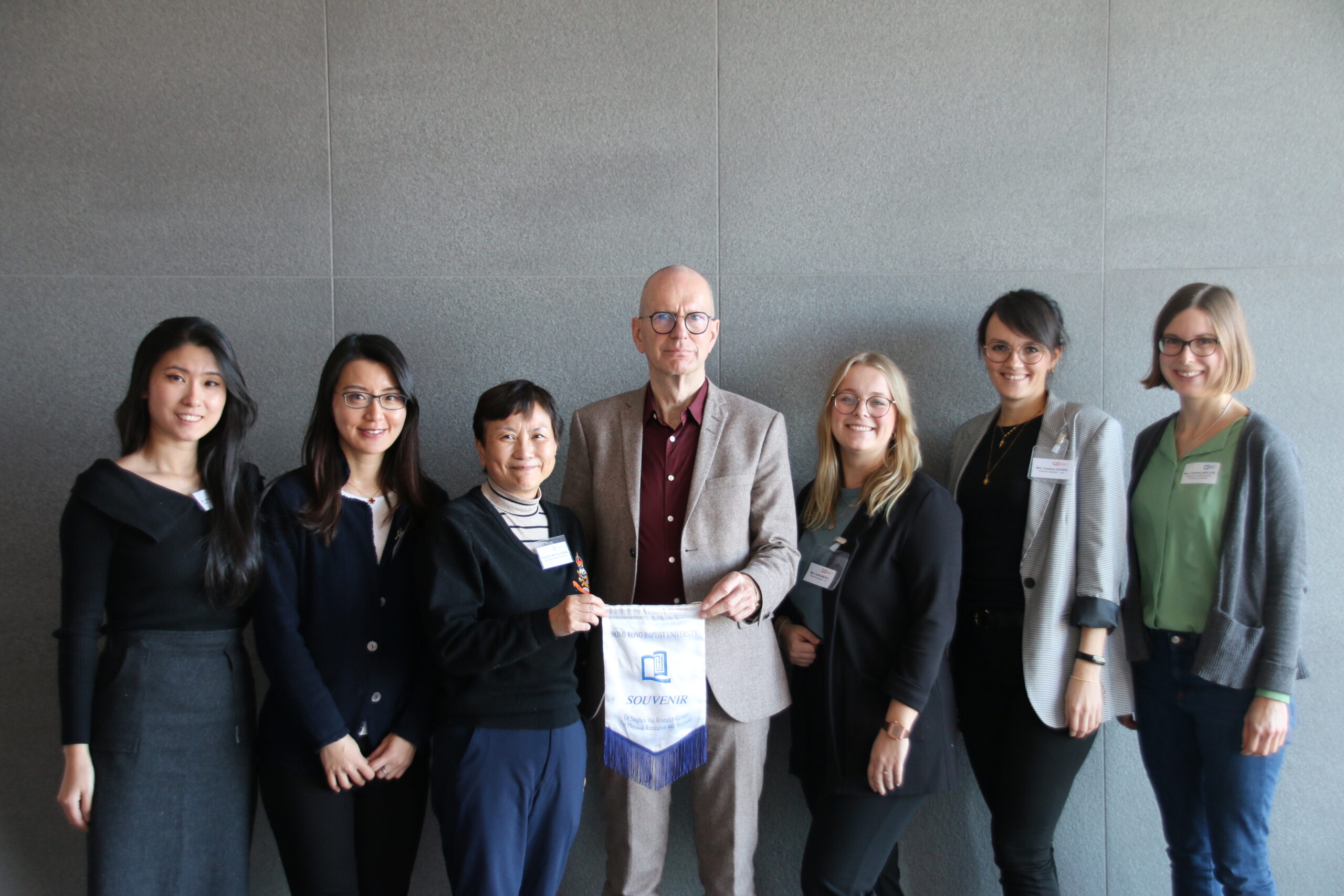 Vor der grauen Wand steht stolz eine Gruppe von sieben Personen, wobei die Person in der Mitte ein „Toastmasters“-Banner hält. Alle lächeln und sind in legerer Business-Kleidung gekleidet, was Selbstvertrauen und Kameradschaft ausstrahlt. Sie repräsentieren die Hochschule Coburg.