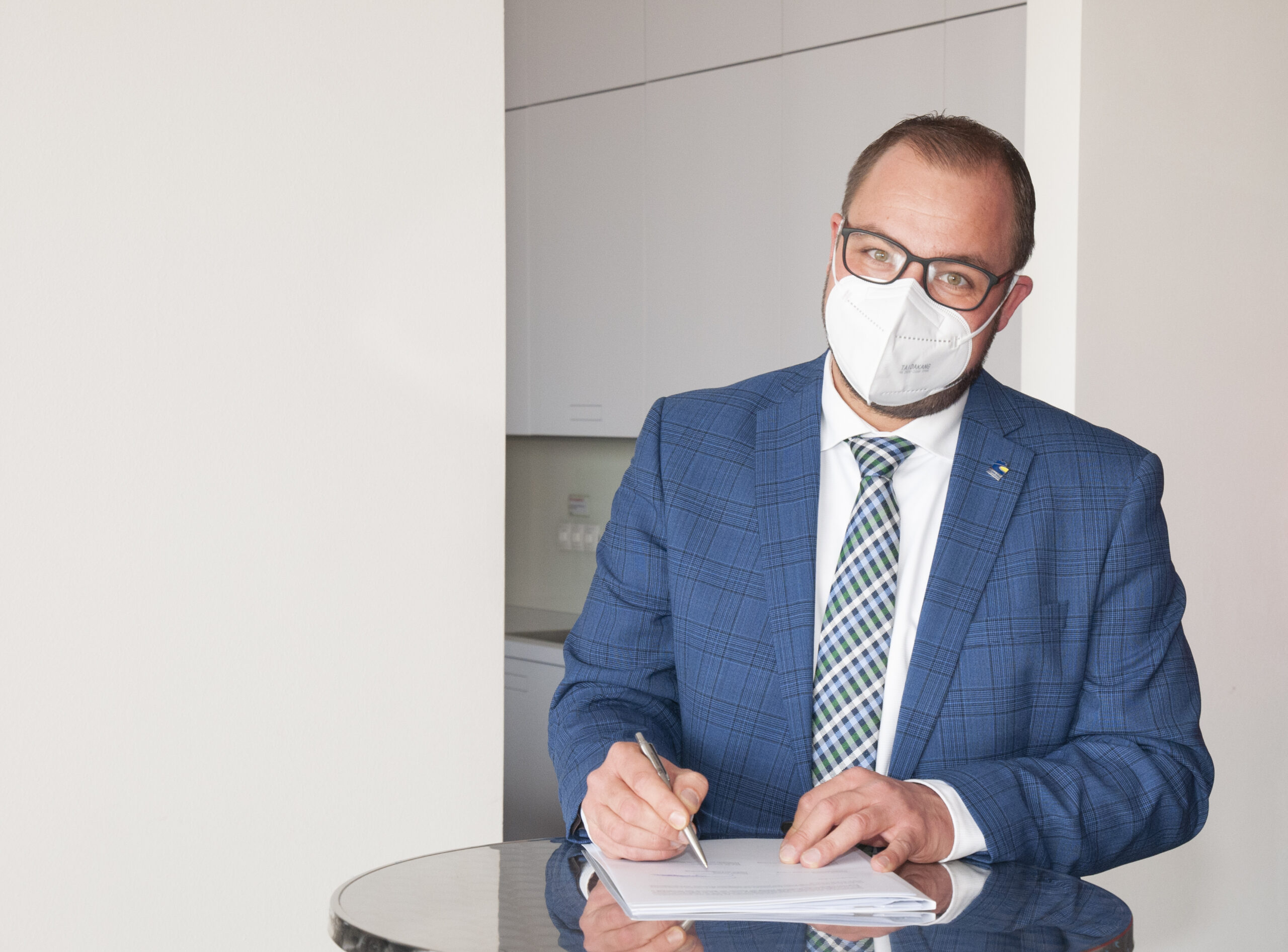 Ein Mann in blauem Anzug und gemusterter Krawatte, der mit der Hochschule Coburg verbunden ist, steht an einem kleinen runden Tisch. Er trägt eine Brille und eine weiße Gesichtsmaske. Er hält einen Stift und scheint auf Papier zu schreiben. Im Hintergrund sind weiße Schränke und eine kahle Wand zu sehen.