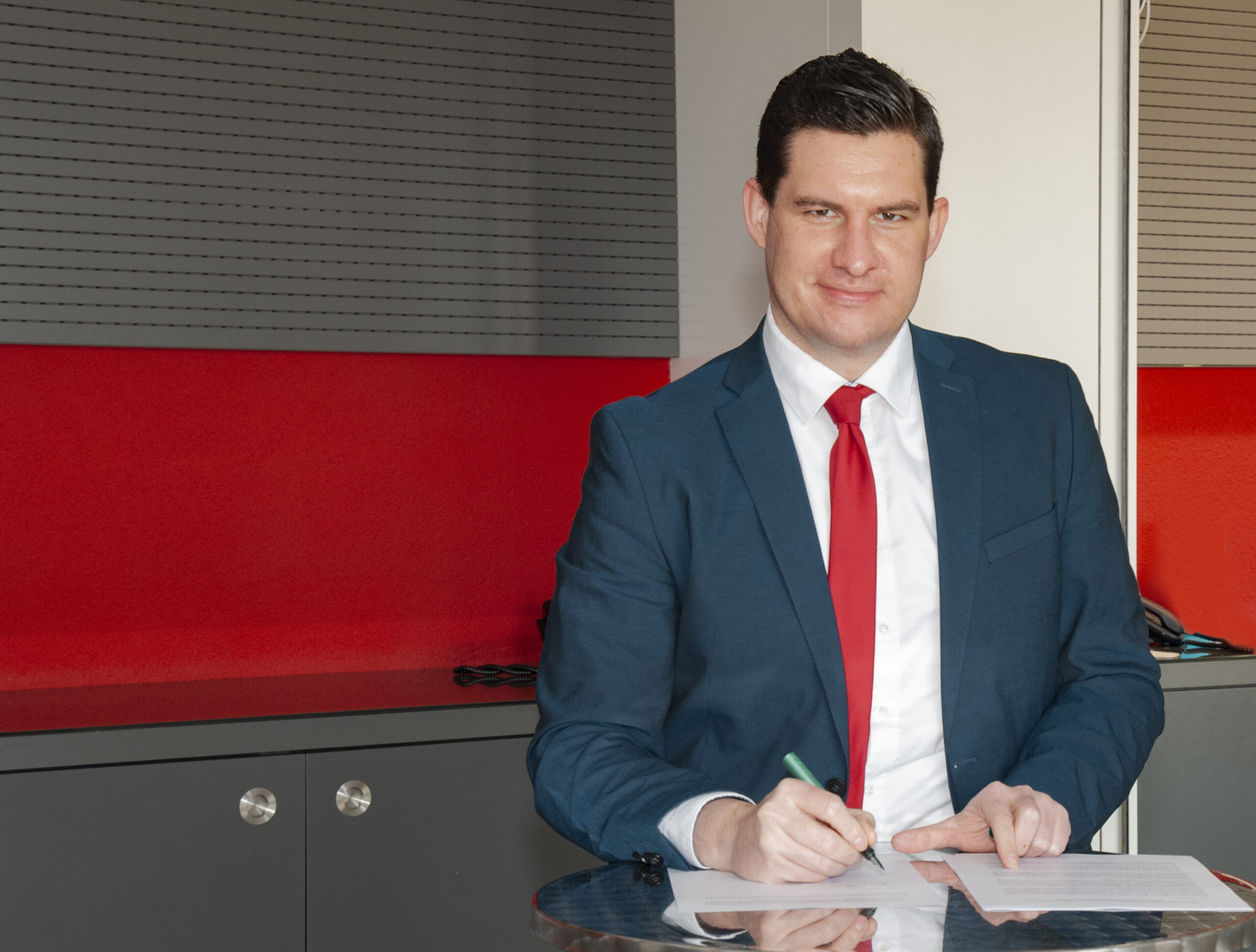 Ein Mann in blauem Anzug und roter Krawatte sitzt mit Stift und Papier in der Hand an einem Glastisch. Die Kulisse ist eine Büroumgebung mit grau-rotem Hintergrund, die an die professionelle Atmosphäre der Hochschule Coburg erinnert.