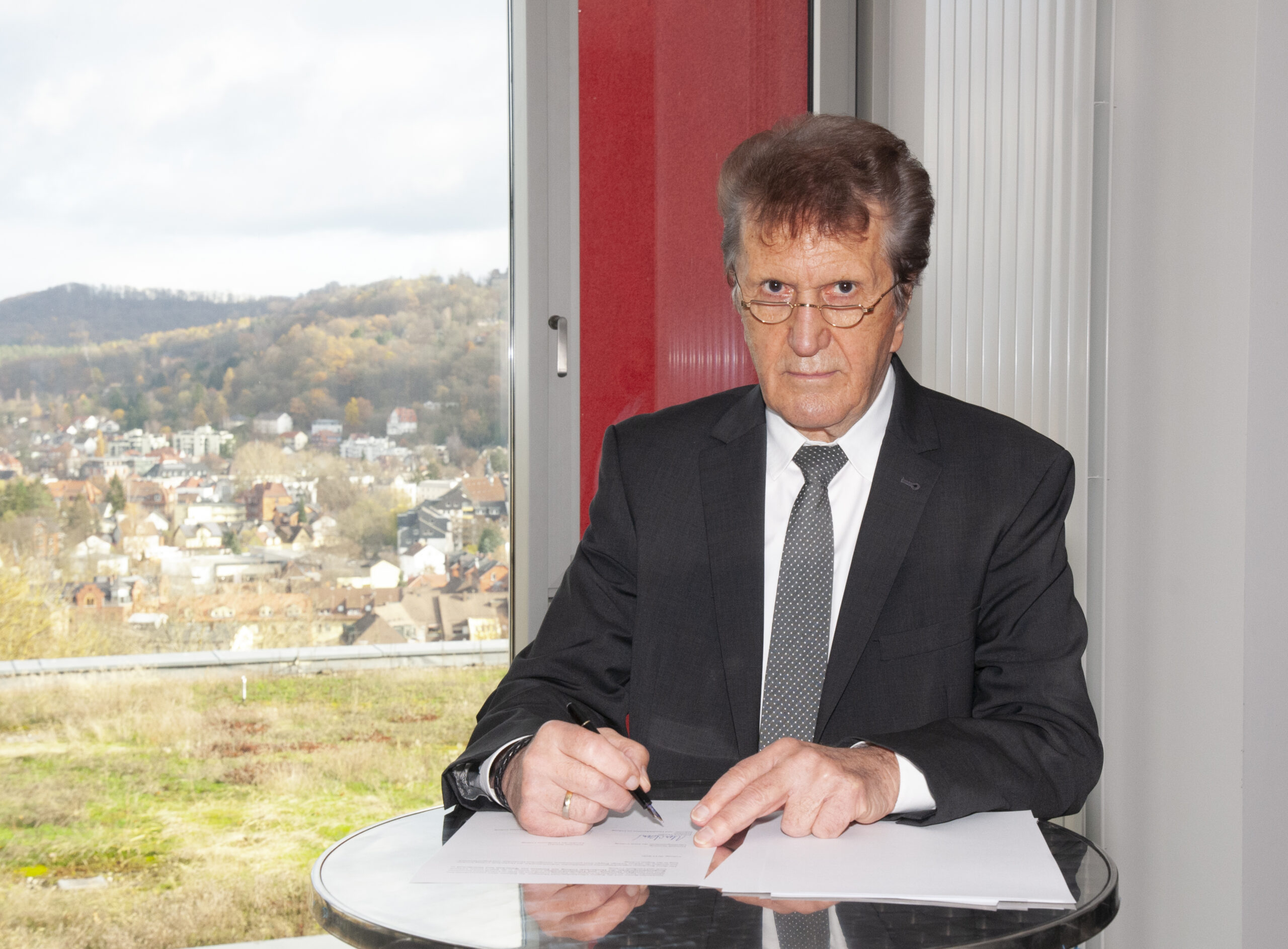 Ein älterer Mann in Anzug und Brille sitzt an einem runden Tisch und unterschreibt ein Dokument der Hochschule Coburg. Durch ein Fenster hinter ihm hat man einen malerischen Blick auf eine Stadt mit Hügeln und Bäumen.