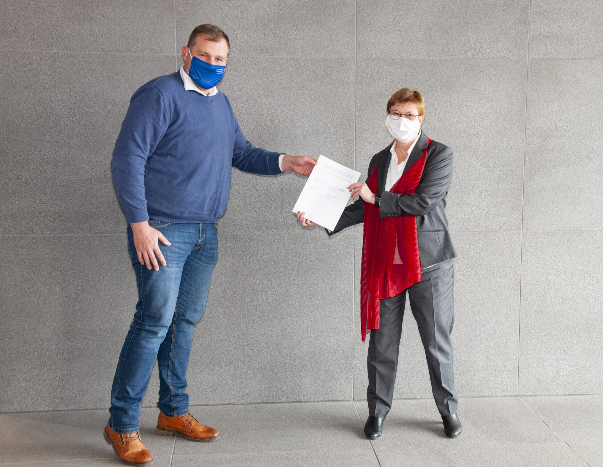 Zwei Menschen mit Masken stehen an einer grauen Wand der Hochschule Coburg. Einer, in blauem Pullover und Jeans, reicht dem anderen, in Anzug und rotem Schal, ein Dokument. Beide halten Abstand, um auf die Pandemie-Vorsichtsmaßnahmen hinzuweisen.