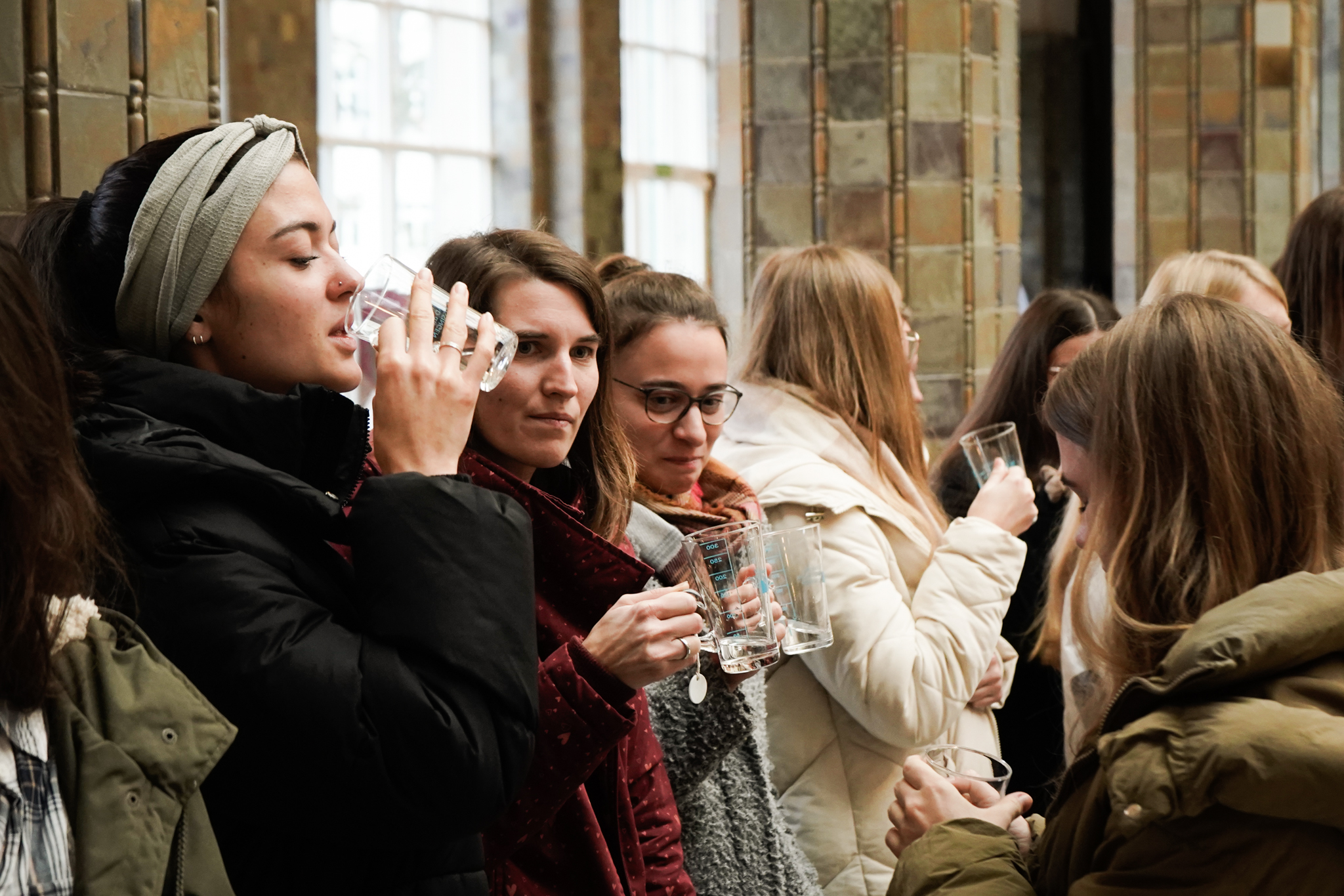 Eine Gruppe winterlich gekleideter Frauen steht in einer Reihe in einem Gebäude mit Backsteinmauern und großen Fenstern der Hochschule Coburg. Sie halten Gläser in den Händen, trinken daraus und scheinen in ein Gespräch vertieft zu sein.