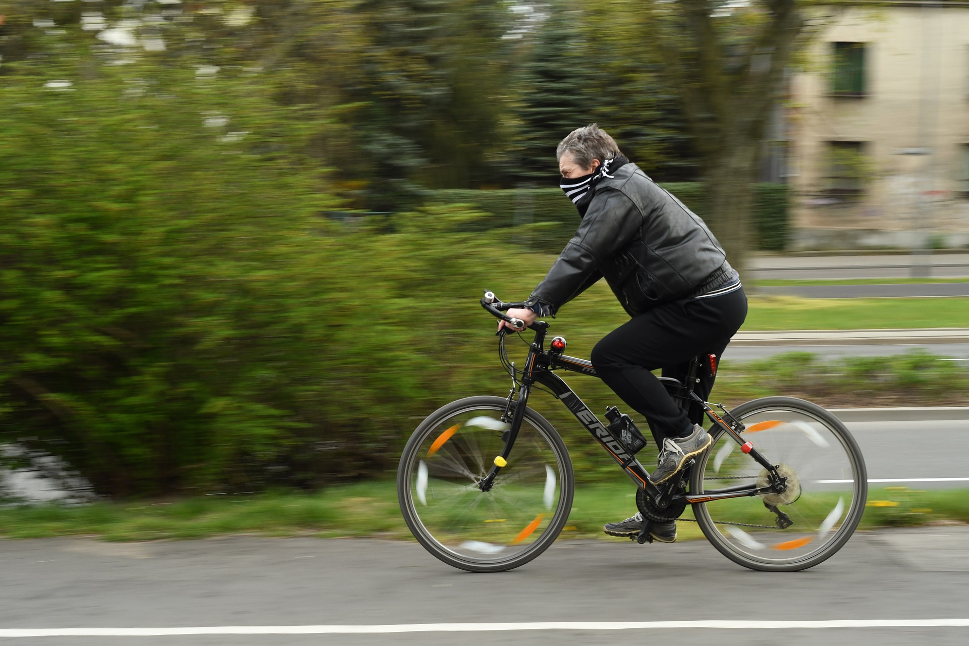 Eine Person mit schwarzer Jacke und Mund-Nasen-Bedeckung fährt auf einem schwarzen Fahrrad zügig an der Hochschule Coburg vorbei. Die grünen Büsche und der unscharfe Hintergrund betonen ihre schnelle Bewegung.