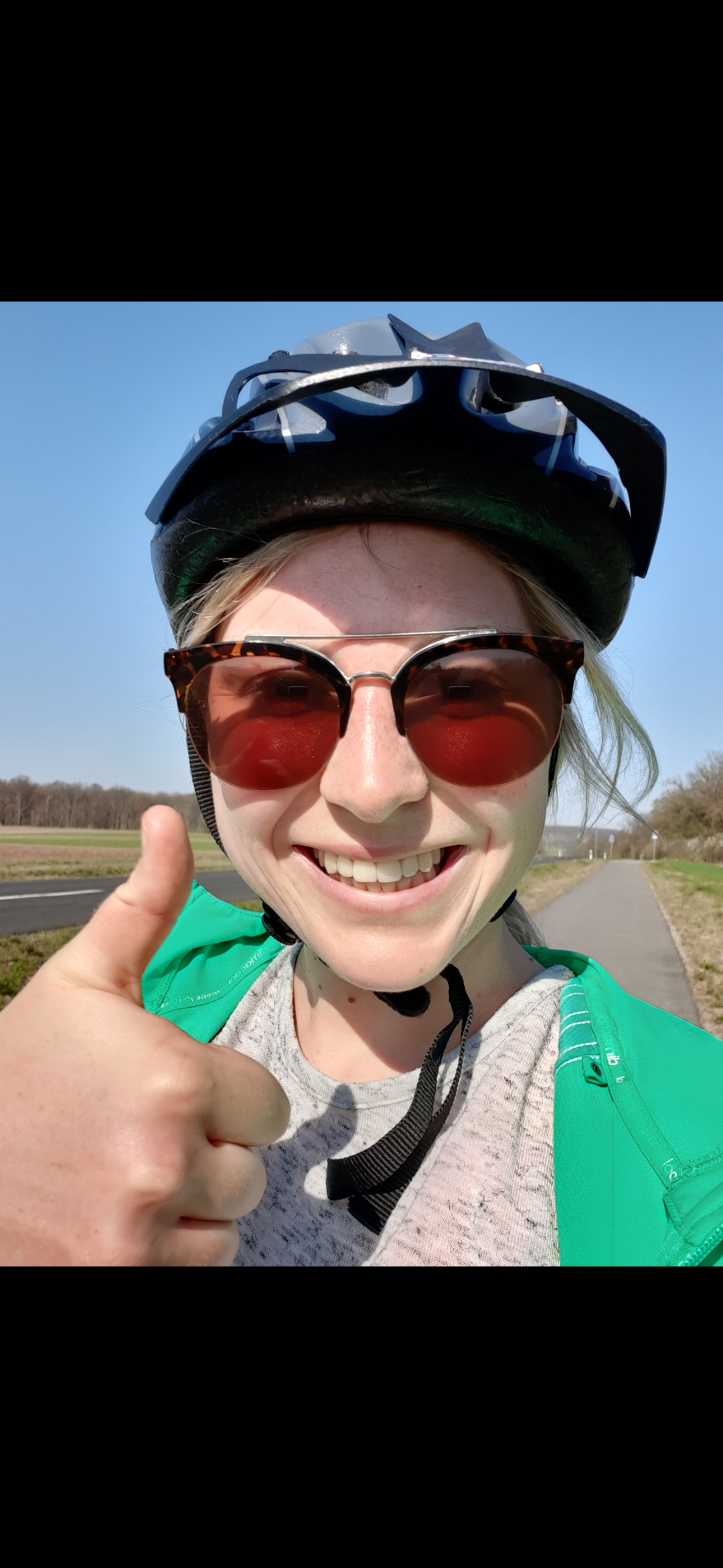 Eine Person mit blauem Fahrradhelm und Sonnenbrille steht auf einem gepflasterten Weg in der Nähe der Hochschule Coburg und lächelt und zeigt den Daumen nach oben. Der Himmel ist klar und blau, im Hintergrund sind einige Bäume zu sehen.