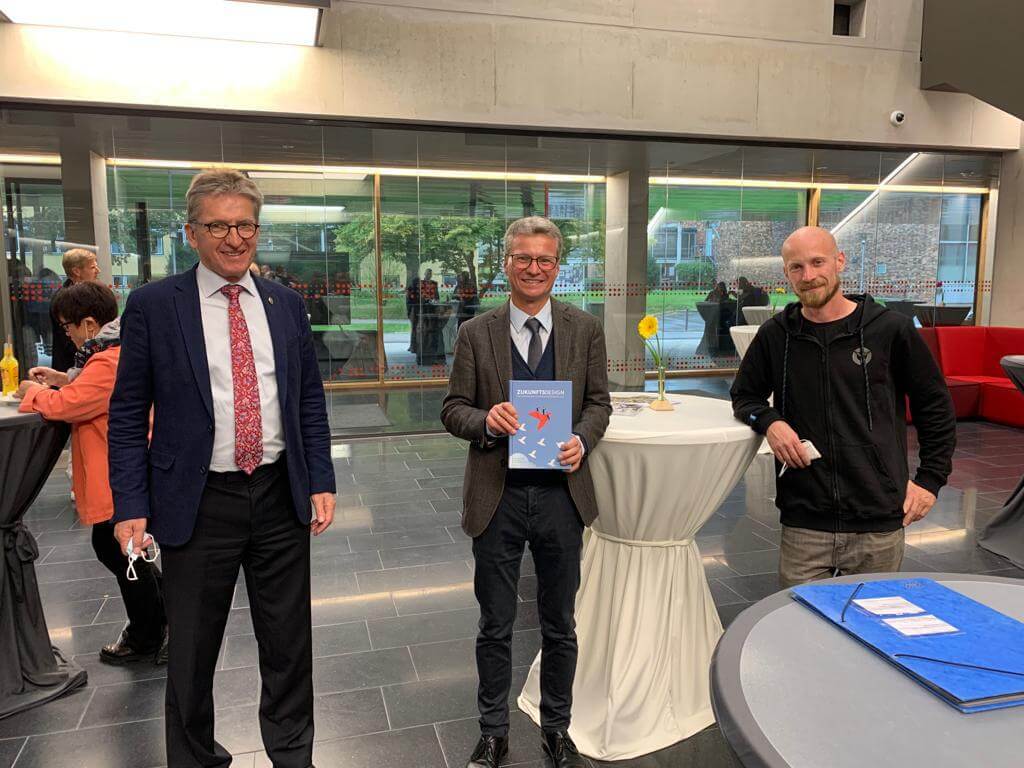 Drei Männer stehen drinnen an einem weiß gedeckten Tisch in der Hochschule Coburg. Der Mann in der Mitte hält ein Buch in der Hand, während sie sich in dem modernen Gebäude mit großen Fenstern versammeln. Im Hintergrund sind ein paar Menschen zu sehen, die zur lebhaften Atmosphäre dieses akademischen Umfelds beitragen.