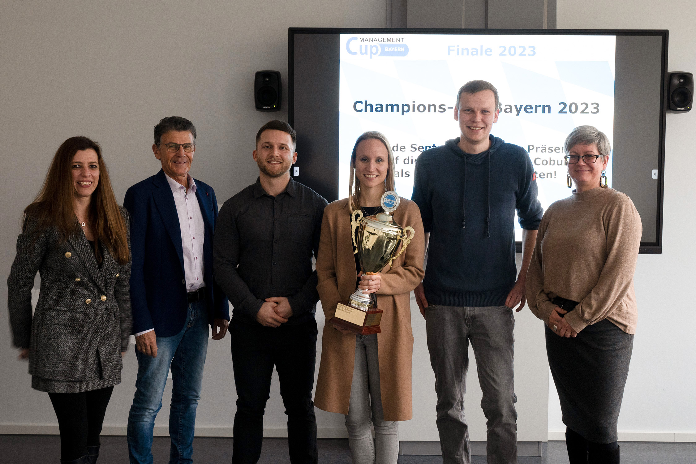 Eine Gruppe von sechs Personen steht gemeinsam vor einem Bildschirm mit der Aufschrift „Champions Bayern 2023“ an der Hochschule Coburg. Eine Person in der Mitte hält stolz eine große Trophäe in den Händen. Sie alle lächeln, tragen eine Mischung aus Freizeit- und Businesskleidung und strahlen Freude und Erfolg aus.