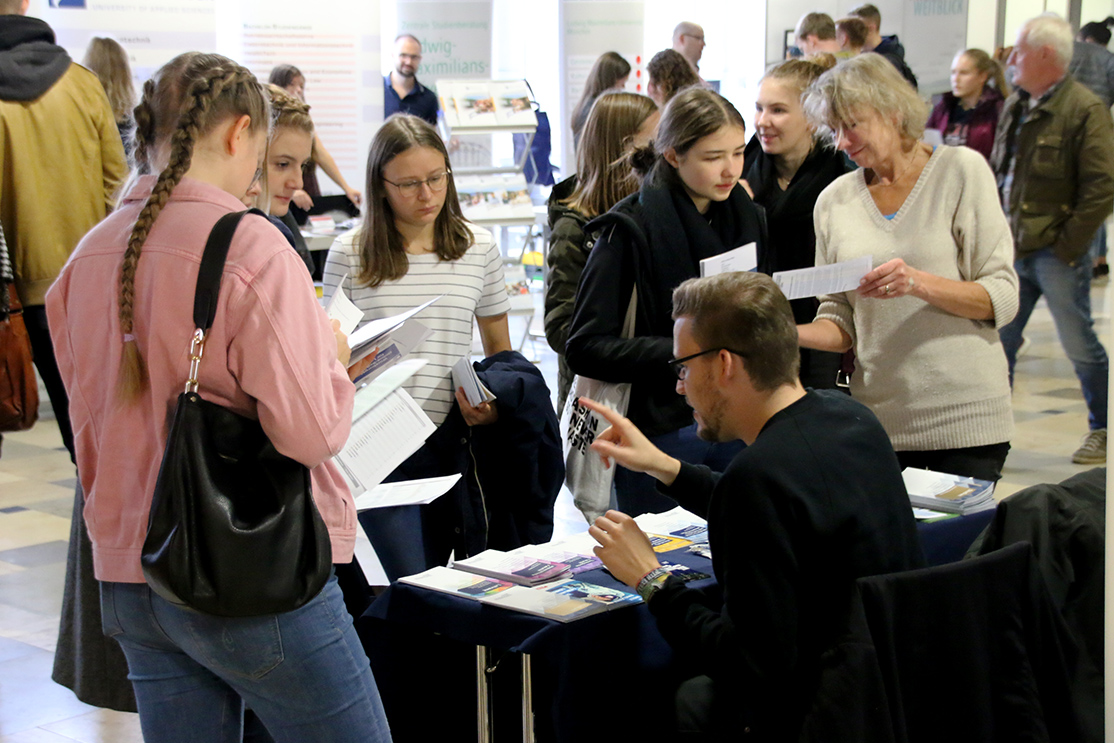 Eine Gruppe von Menschen versammelt sich um einen Stand bei der Veranstaltung der Hochschule Coburg, wo ein Mann an einem Tisch sitzt und Broschüren verteilt. Eine Frau in der Gruppe hält einen Vortrag und führt lebhafte Diskussionen. Einige sind fasziniert von den ausgestellten Materialien. Die Veranstaltung findet eindeutig in Innenräumen statt.