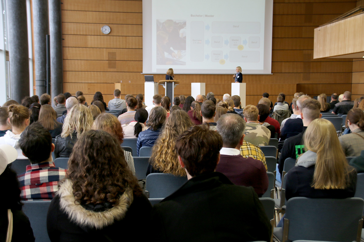 Der Hörsaal der Hochschule Coburg ist voller sitzender Zuhörer, die vor der Bühne sitzen. Zwei Vortragende stehen an Rednern vor einer großen Leinwand, auf der eine Folie gezeigt wird. Das bunt gemischte Publikum präsentiert eine Reihe von Frisuren und Kleidungsstilen, die die lebendige Campus-Gemeinschaft widerspiegeln.