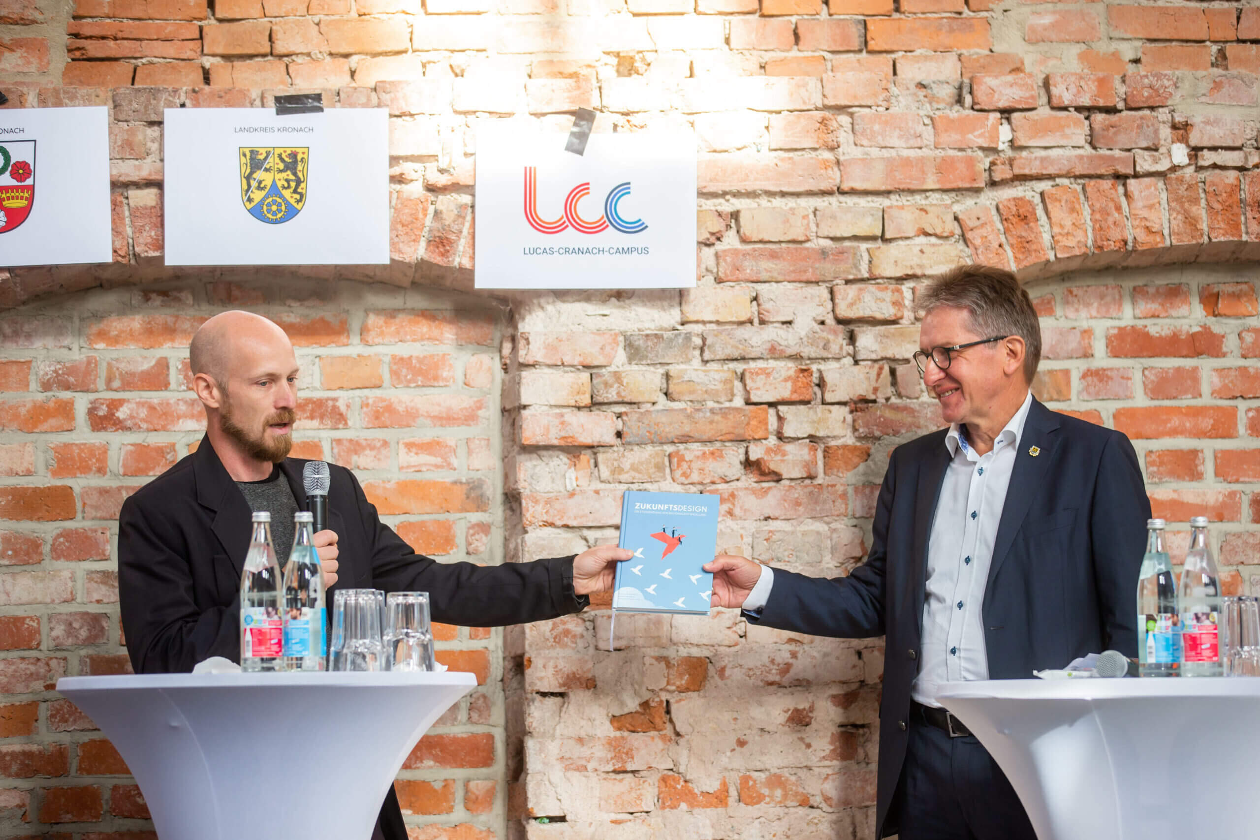 Zwei Männer stehen an runden Tischen vor einer Backsteinwand. Einer hält ein Mikrofon in der Hand und präsentiert dem anderen ein Buch. Beide tragen Anzüge. An der Wand hinter ihnen hängen Banner mit Logos der Hochschule Coburg, auf den Tischen stehen Wasserflaschen.