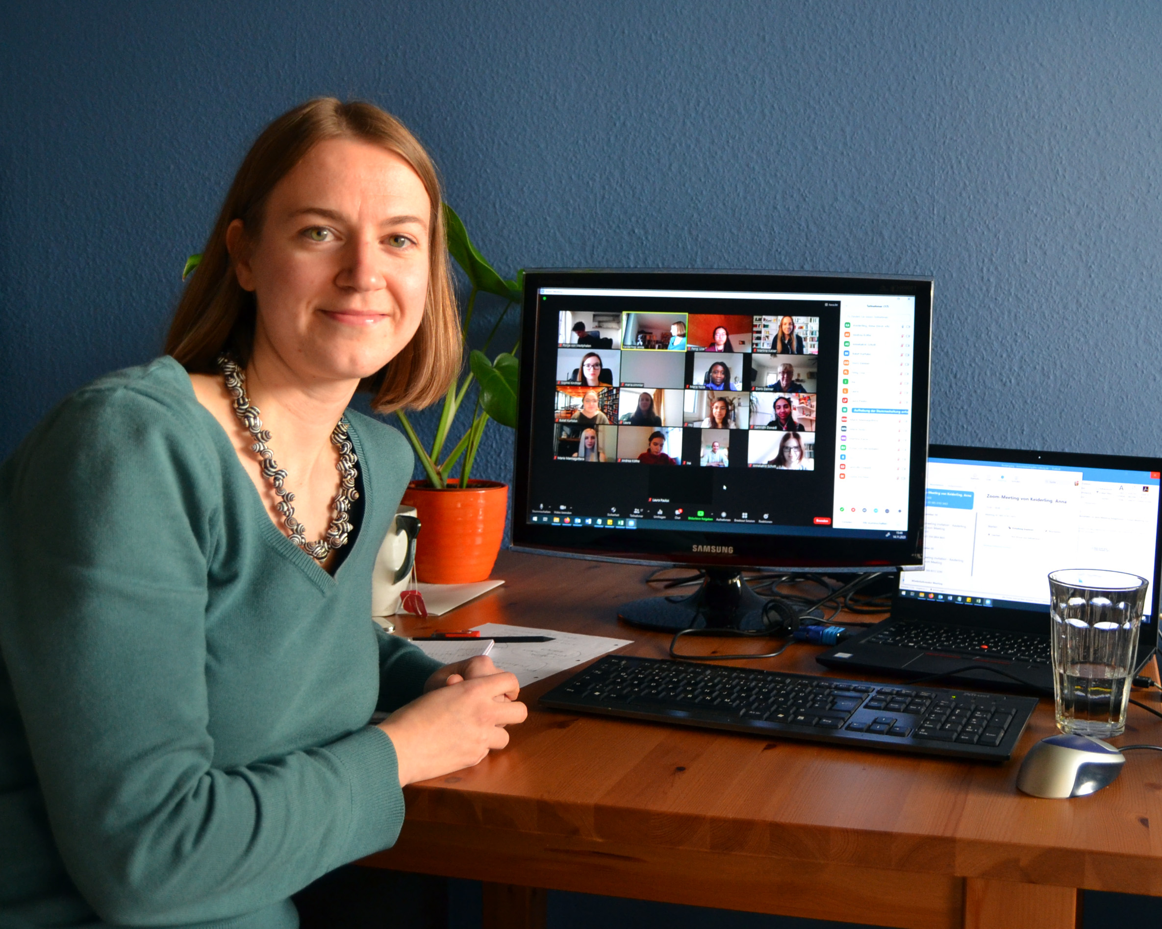 Eine Frau in einem grünen Pullover sitzt an einem Holzschreibtisch und lächelt in die Kamera. Sie nimmt an einer Videokonferenz der Hochschule Coburg teil, die auf einem Computermonitor angezeigt wird. Auf dem Schreibtisch stehen außerdem ein Laptop, ein Glas Wasser und eine Topfpflanze.