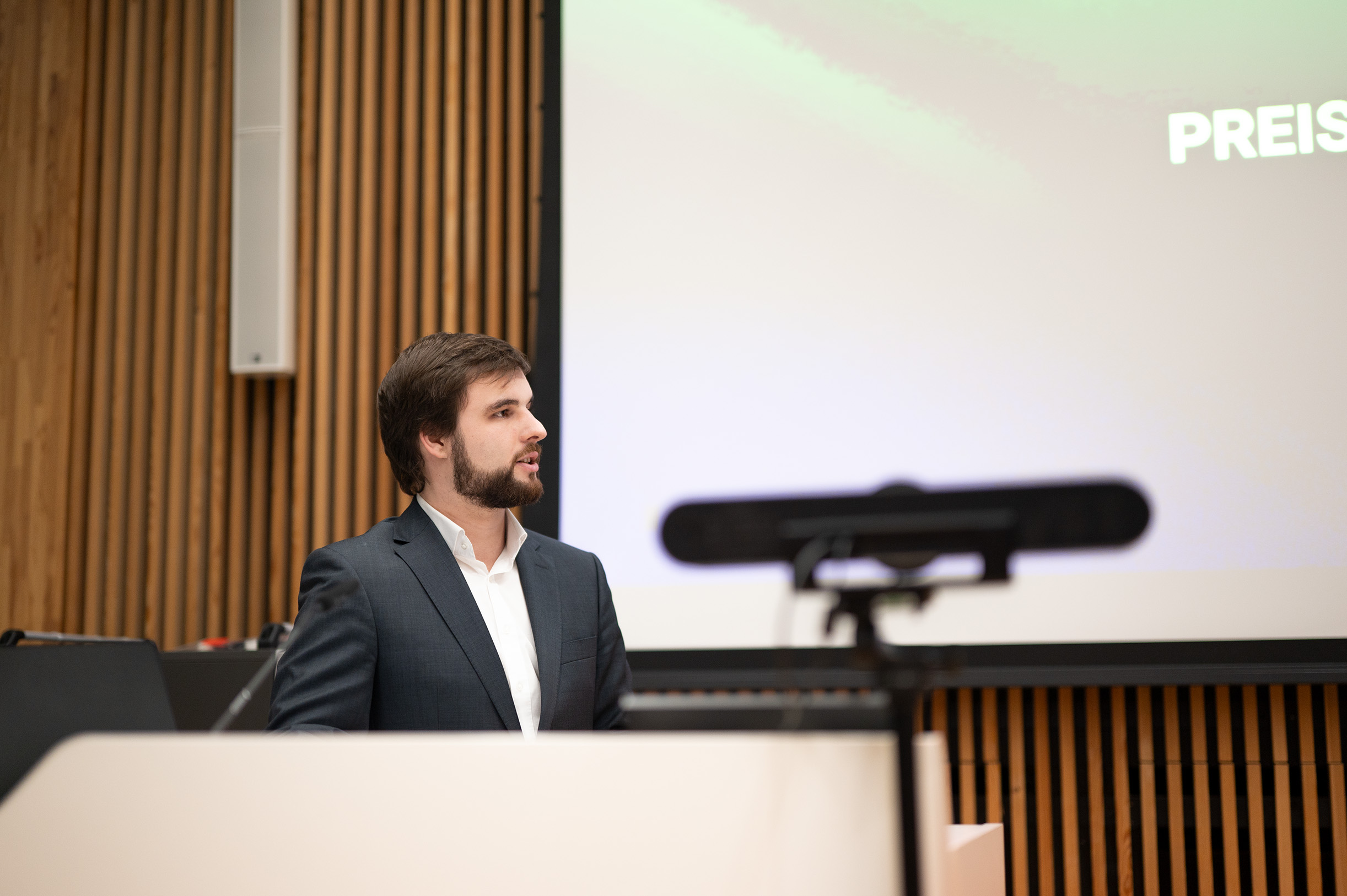 Ein Mann im Anzug steht an einem Podium und spricht vor einem großen Bildschirm, auf dem das Wort „PREIS“ zu sehen ist. Die Kulisse, möglicherweise ein Teil der Hochschule Coburg, sieht aus wie ein Konferenz- oder Hörsaal mit Holzvertäfelung an den Wänden. Im Vordergrund ist ein Mikrofon zu sehen.