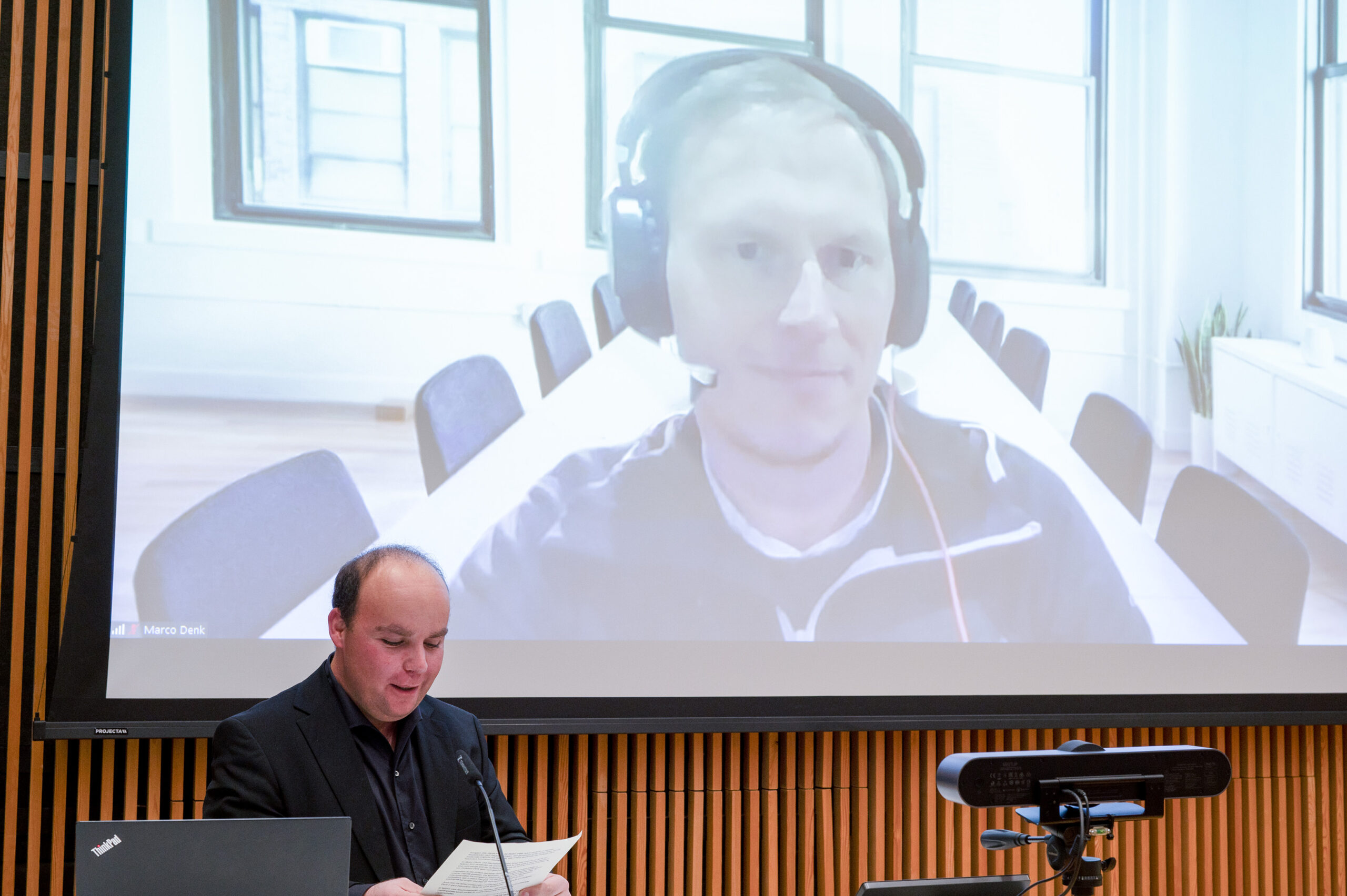 Eine Person steht an einem Podium und liest aus einem Artikel vor, während ein großer Bildschirm dahinter eine andere Person in einem Videoanruf zeigt. Der moderne Raum, der an den Stil der Hochschule Coburg erinnert, verfügt über Holzwandpaneele und große Fenster, die einen beeindruckenden virtuellen Hintergrund schaffen.