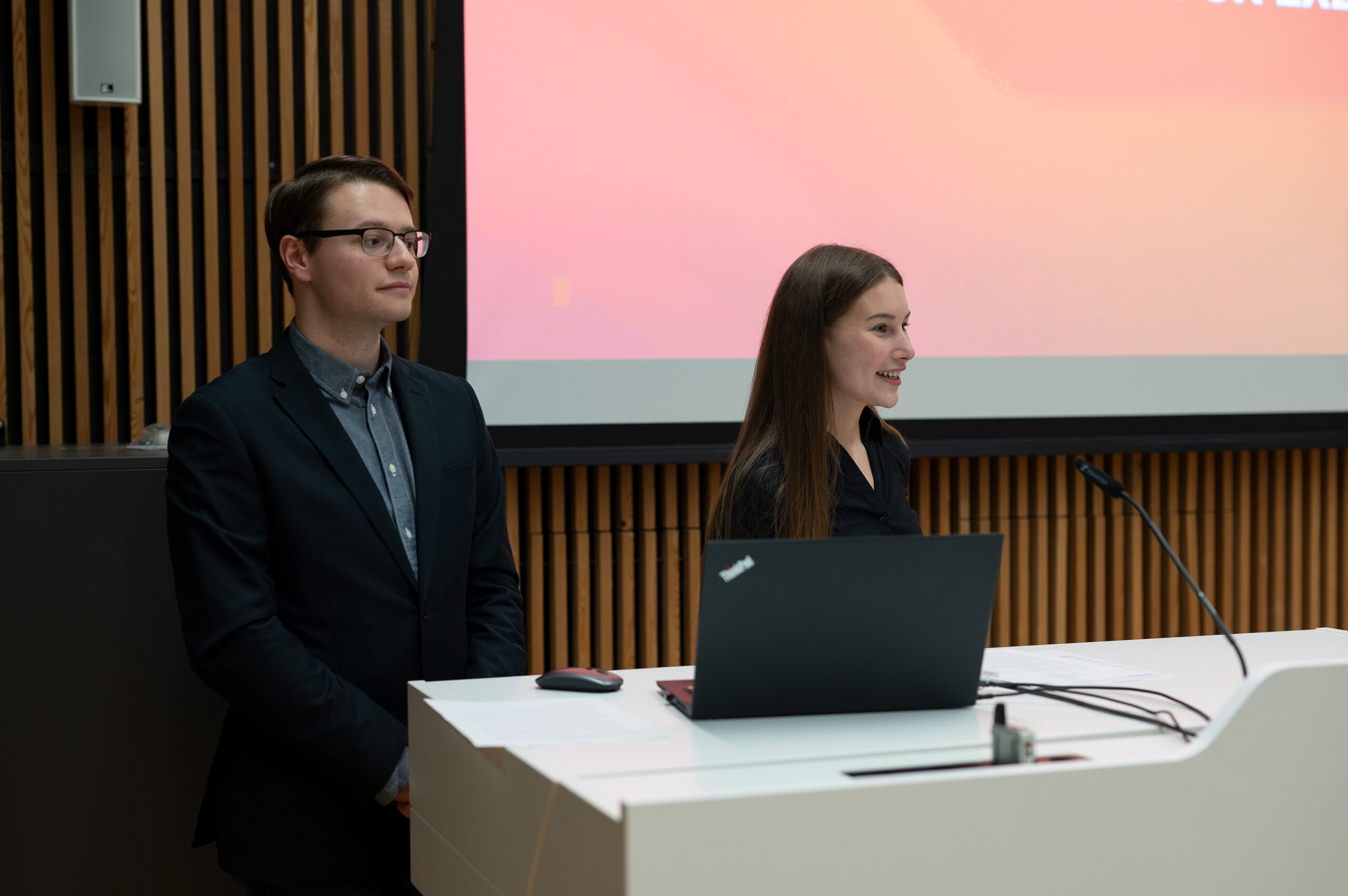 Zwei Personen stehen mit einem Laptop an einem Podium und präsentieren vor einem großen Bildschirm mit Farbverlauf. Die Kulisse, vermutlich ein Konferenz- oder Hörsaal mit Holzvertäfelung, spiegelt das akademische Ambiente der Hochschule Coburg wider.