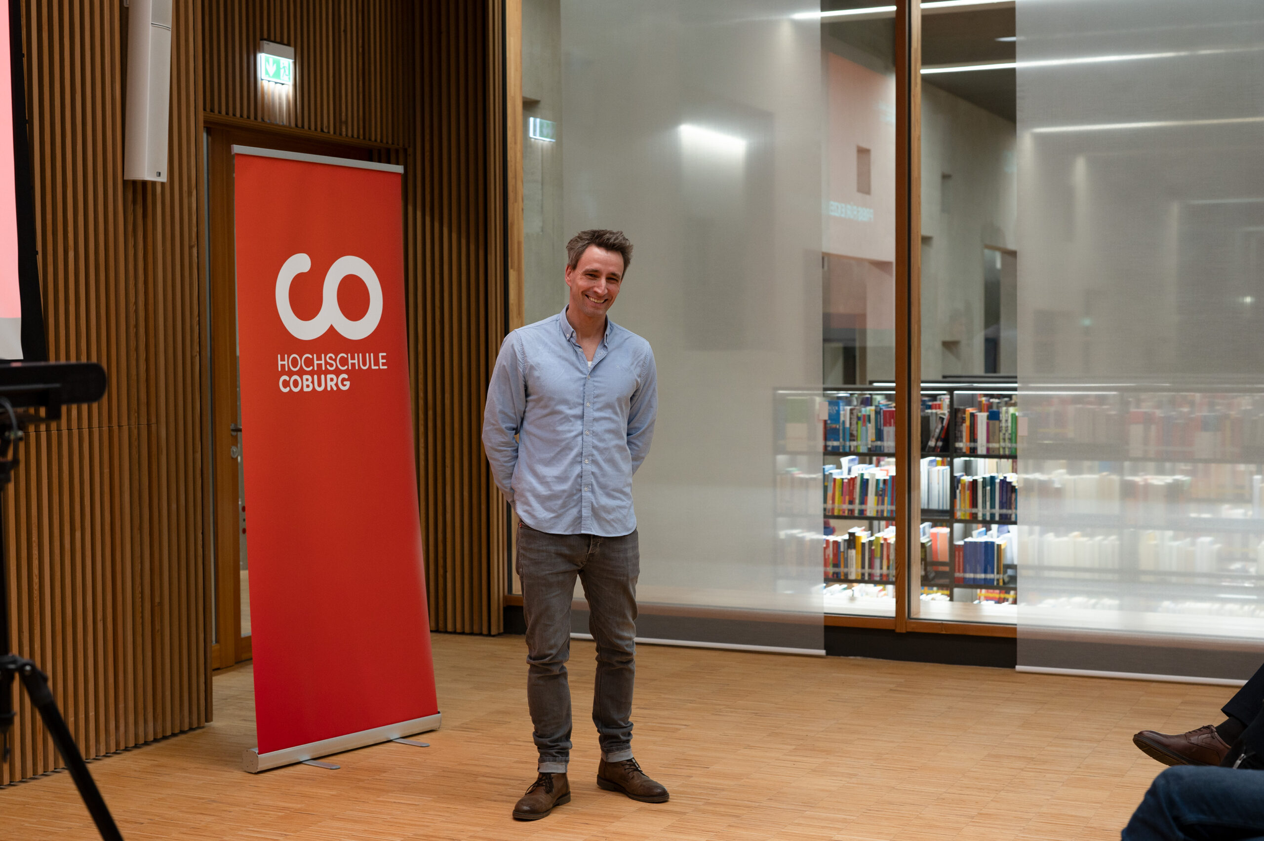 Eine Person steht lächelnd vor einem roten Banner der Hochschule Coburg in einem modernen, hellen Raum. Hinter der Glaswand sind Bücher zu sehen, der Holzboden verleiht der Szene Wärme.