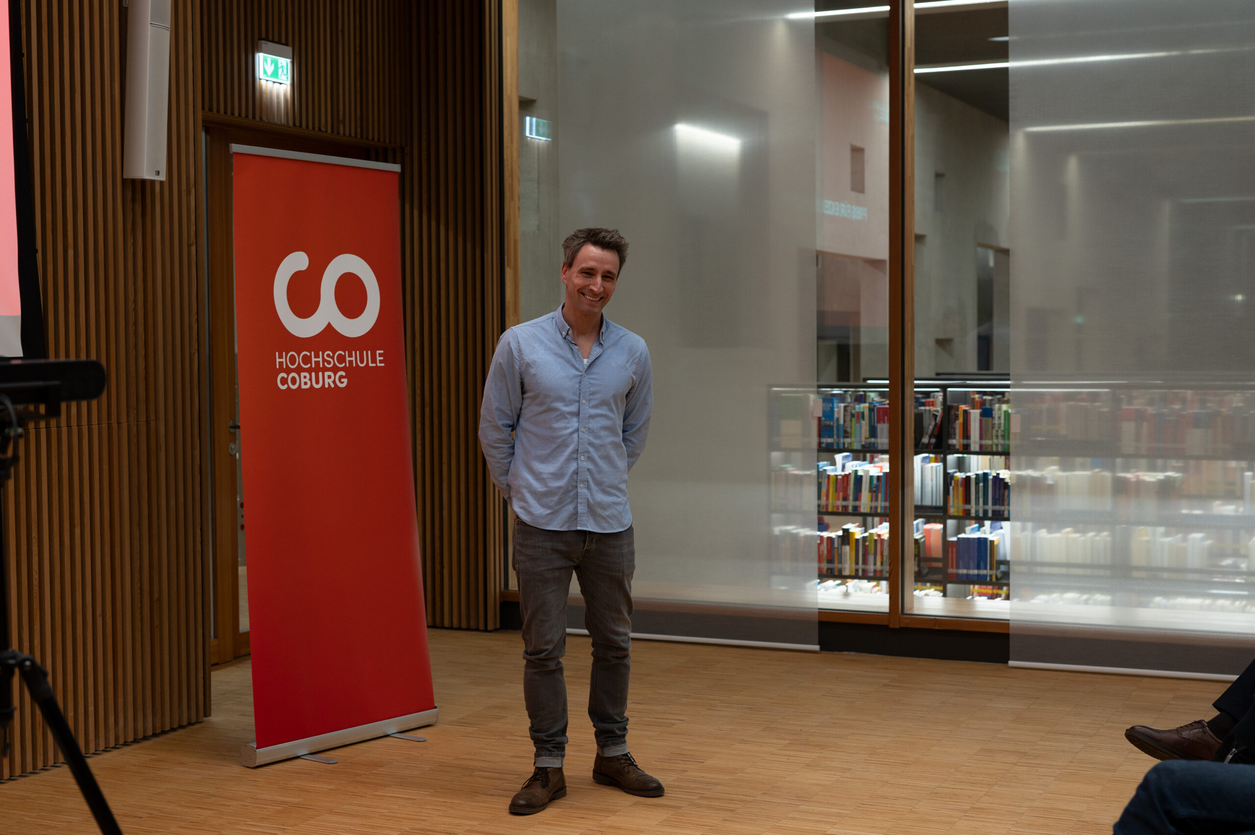 In einem modernen Raum mit Holzvertäfelung und großen Fenstern steht eine lächelnde Person und hält stolz ein rotes „Hochschule Coburg“-Banner in die Höhe. Dahinter tragen Regale voller Bücher zur akademischen Atmosphäre bei.