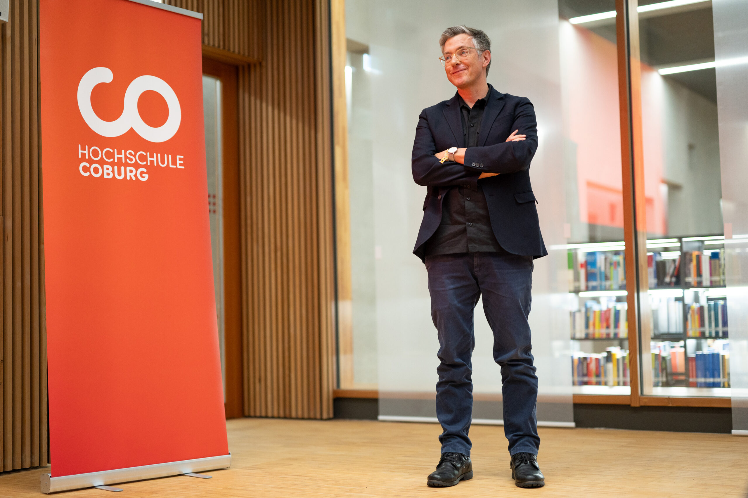 Eine Person in dunkler Jacke und Jeans steht mit verschränkten Armen in einer Bibliothek und strahlt Selbstbewusstsein aus. Links davon ist ein rotes Banner mit der Aufschrift „Hochschule Coburg“ zu sehen, während im Hintergrund Bücherregale stehen, die eine akademische Atmosphäre schaffen.