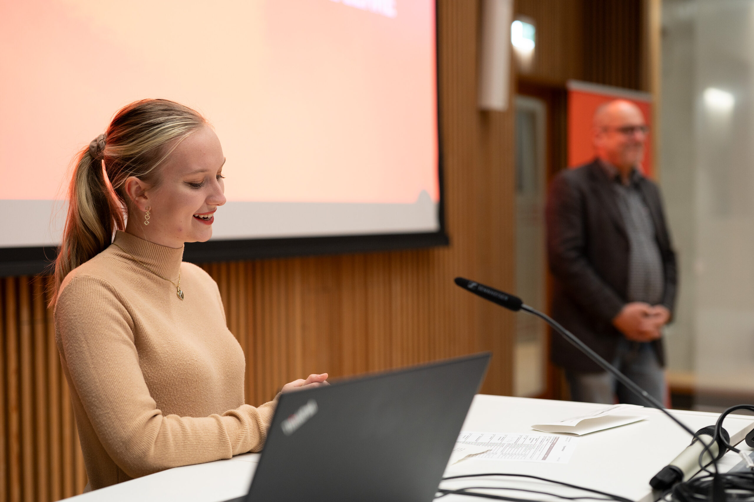 Eine Frau im beigen Pullover spricht an einem Podium mit einem Laptop vor einer Projektionsleinwand, im Hintergrund steht ein Mann im Jackett. Sie sind im modernen Konferenzraum der Hochschule Coburg mit seinen glatten Holzwänden versammelt.