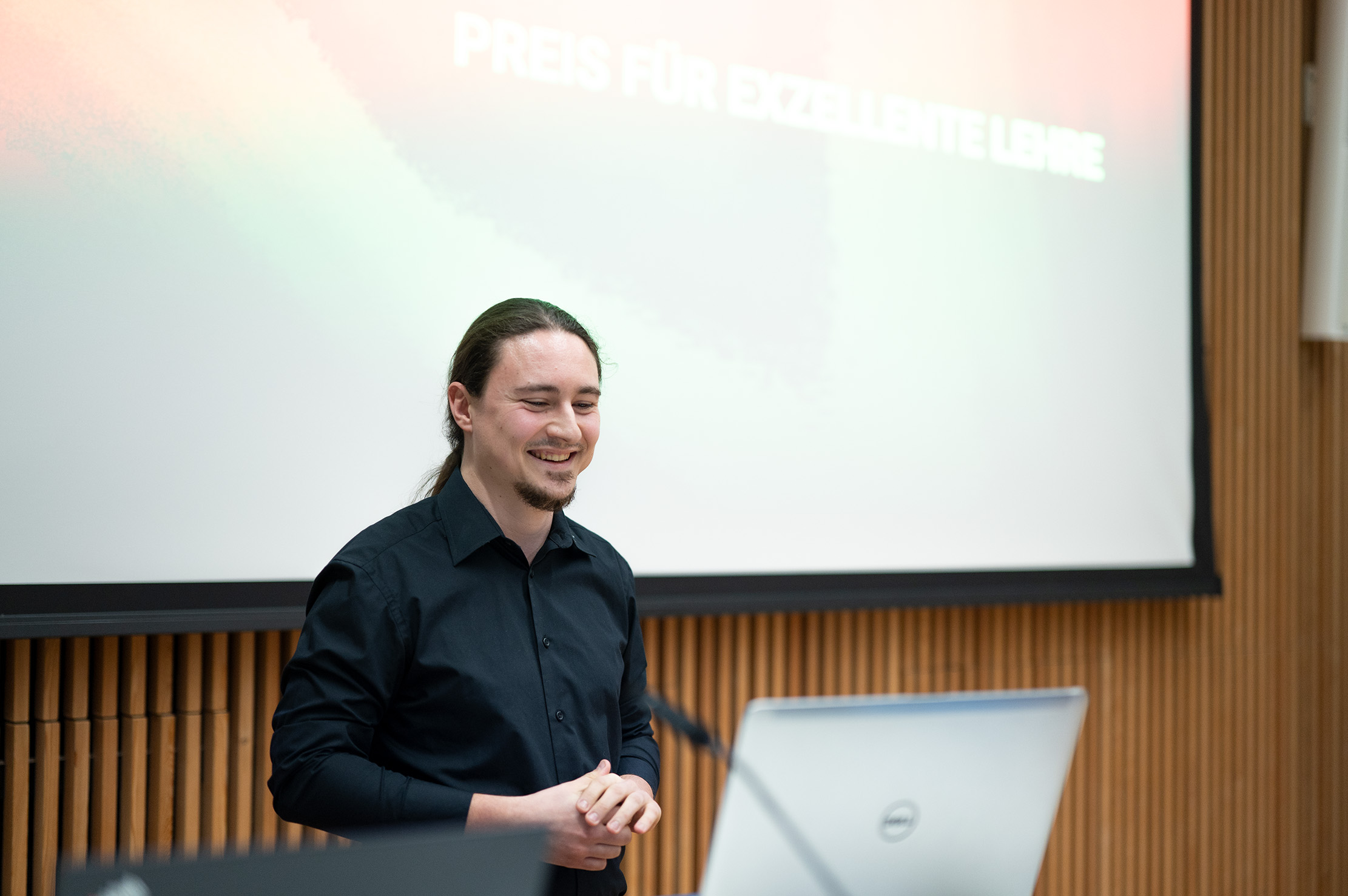 Eine Person mit langen Haaren und Bart, bekleidet mit einem schwarzen Hemd, steht lächelnd vor einem Podium mit einem Dell-Laptop an der Hochschule Coburg. Dahinter ist ein unscharfer Präsentationsbildschirm mit der deutschen Aufschrift „PREIS FÜR EXZELLENTE LEHRE“ zu sehen.