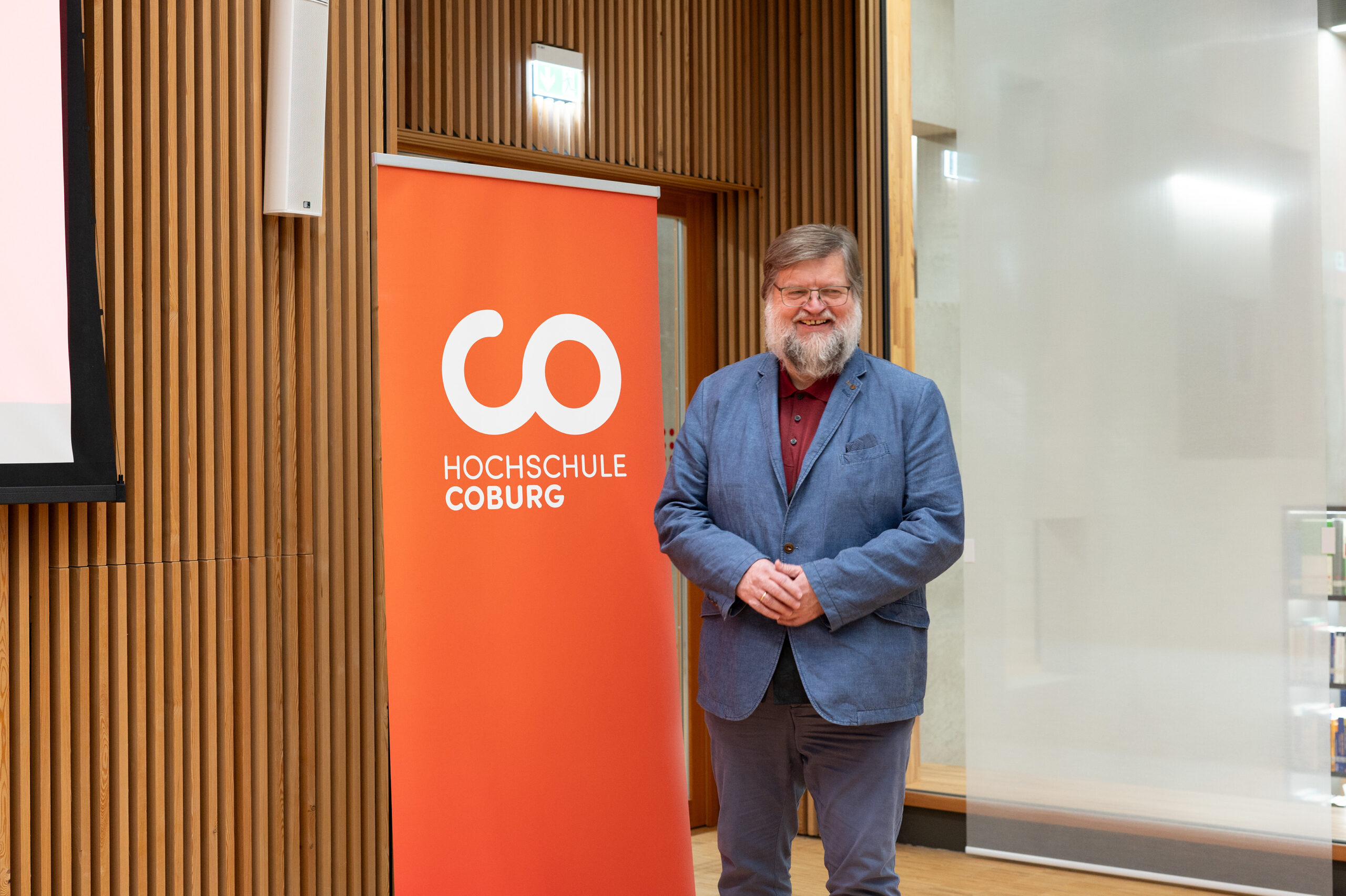 Ein bärtiger Mann im blauen Anzug steht lächelnd neben einem orangefarbenen Banner mit der Aufschrift „Hochschule Coburg“ in strahlend weißem Text, das Ganze vor der Kulisse eines modernen holzgetäfelten Raums.