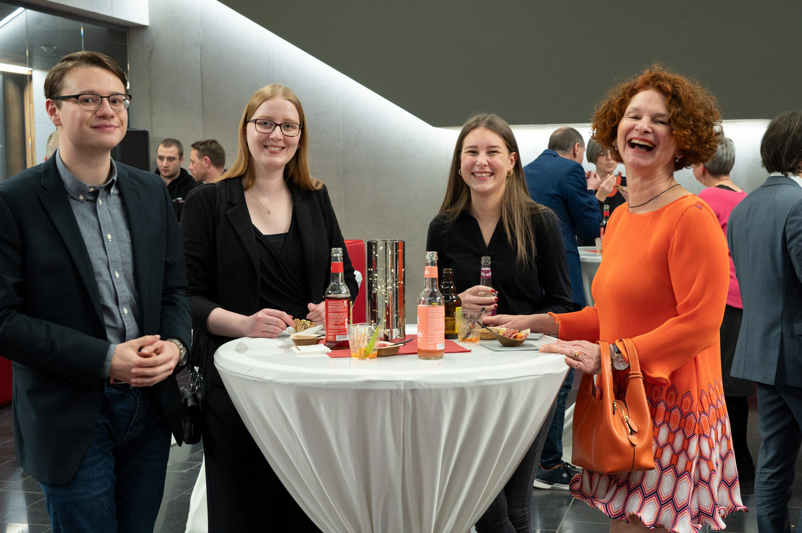 Eine Gruppe von vier Personen steht bei einer Veranstaltung der Hochschule Coburg mit Getränken und Snacks um einen Stehtisch. Sie sind halbformell gekleidet, lächeln und unterhalten sich miteinander. Im Hintergrund sind andere Teilnehmer zu sehen, die sich in einem modernen Innenbereich unterhalten und zur lebhaften Atmosphäre beitragen.