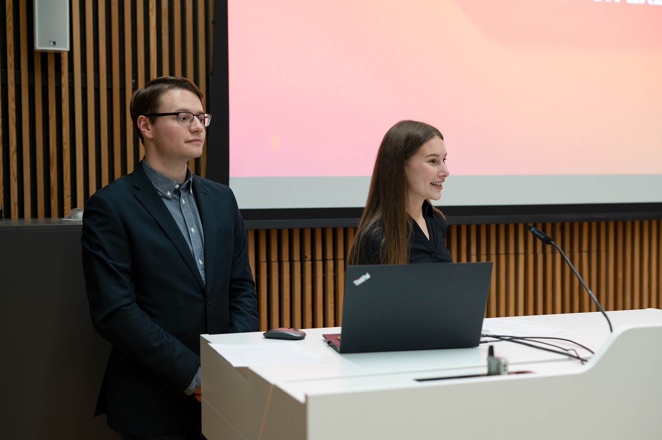 Ein Mann und eine Frau von der Hochschule Coburg stehen an einem Podium und halten einen Vortrag. Der Mann trägt eine Brille und einen Anzug, die Frau ein dunkles Oberteil. Auf dem Podium steht ein aufgeklappter Laptop, im Hintergrund zeigt ein großer Bildschirm einen Farbverlauf.