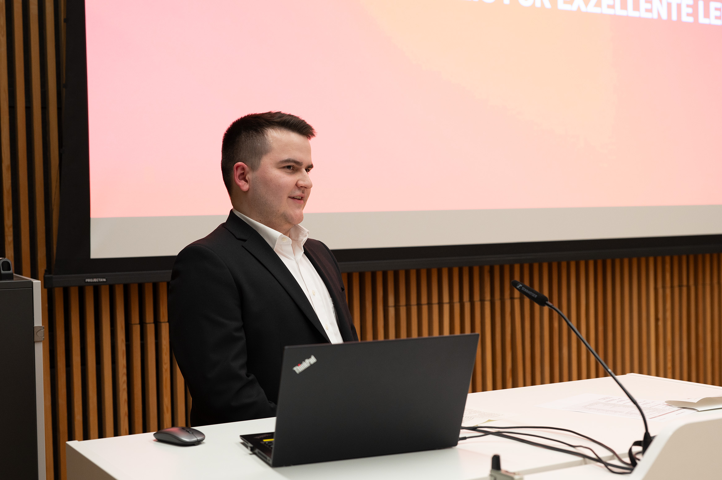 Eine Person im Anzug spricht an einem Podium mit einem Mikrofon und einem Laptop vor sich und vertritt die Hochschule Coburg. Im Hintergrund wird eine rote Präsentationsfolie angezeigt, die auf eine formelle Veranstaltung oder Konferenz hindeutet.