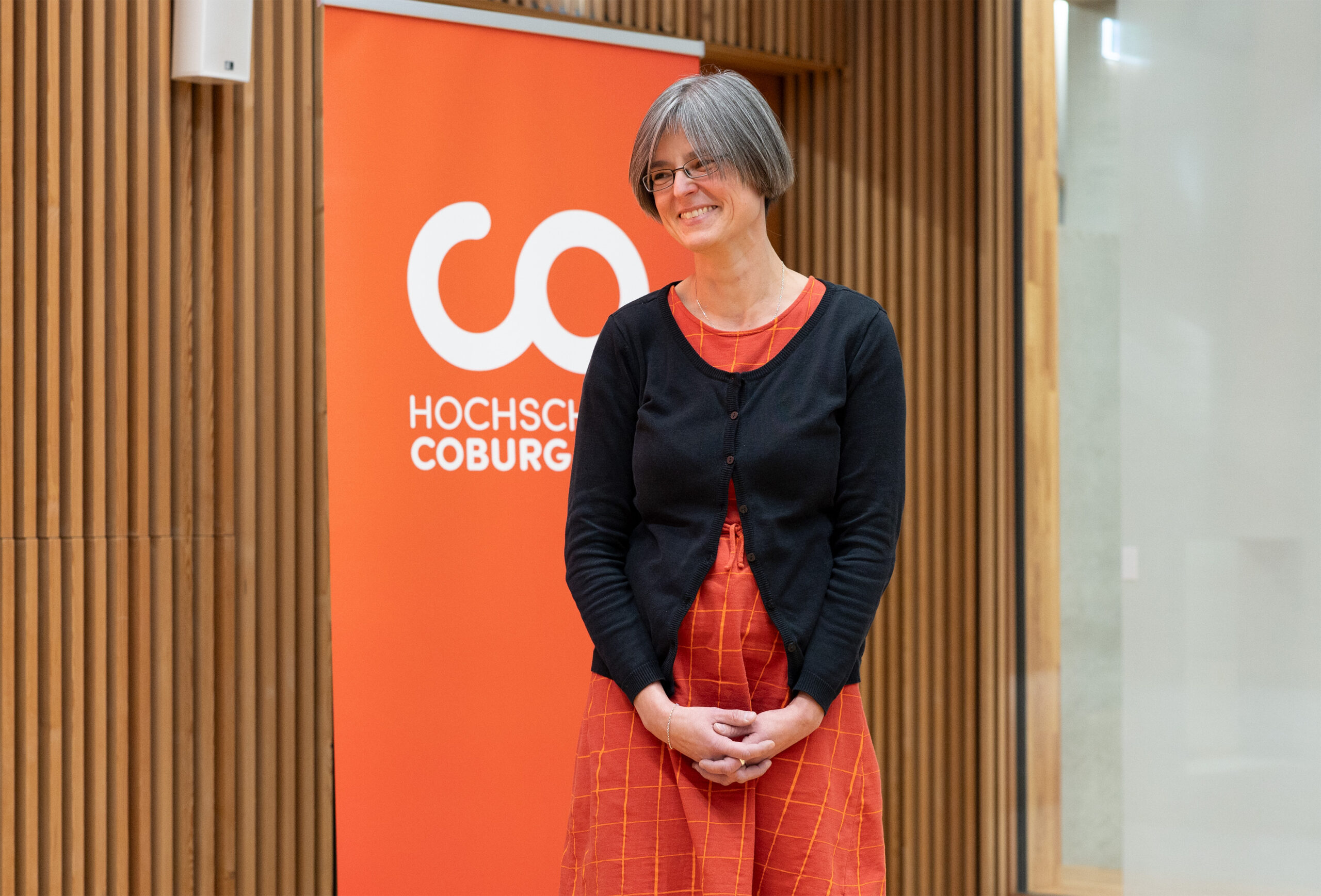 Eine Person mit kurzen grauen Haaren, Brille, schwarzer Strickjacke und orangefarbenem Kleid steht vor einem orangefarbenen Schild mit dem Logo der „HOCHSCHULE COBURG“. Im Hintergrund ist eine markante Holzvertäfelung zu sehen.