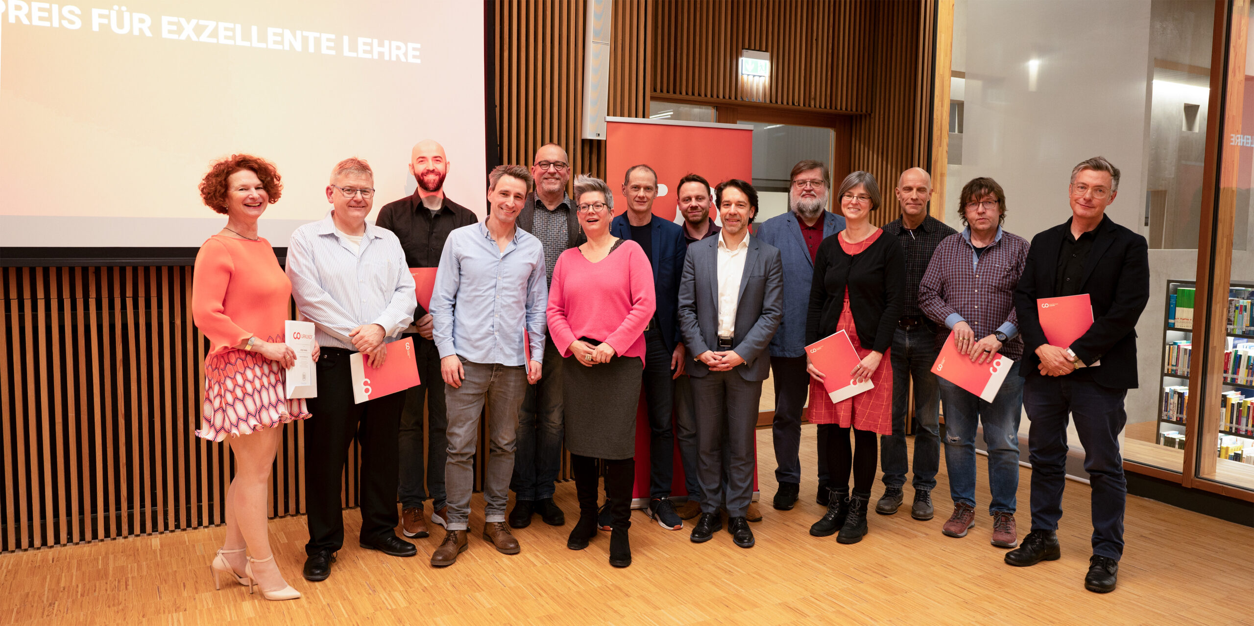 Eine Gruppe von Menschen steht auf dem Holzboden eines Raums an der Hochschule Coburg. Vor ihr steht ein Bildschirm mit der Aufschrift „Preis für exzellente Lehre“. Einige halten rote Umschläge in den Händen, was darauf schließen lässt, dass sie an einer Preisverleihung teilnehmen.