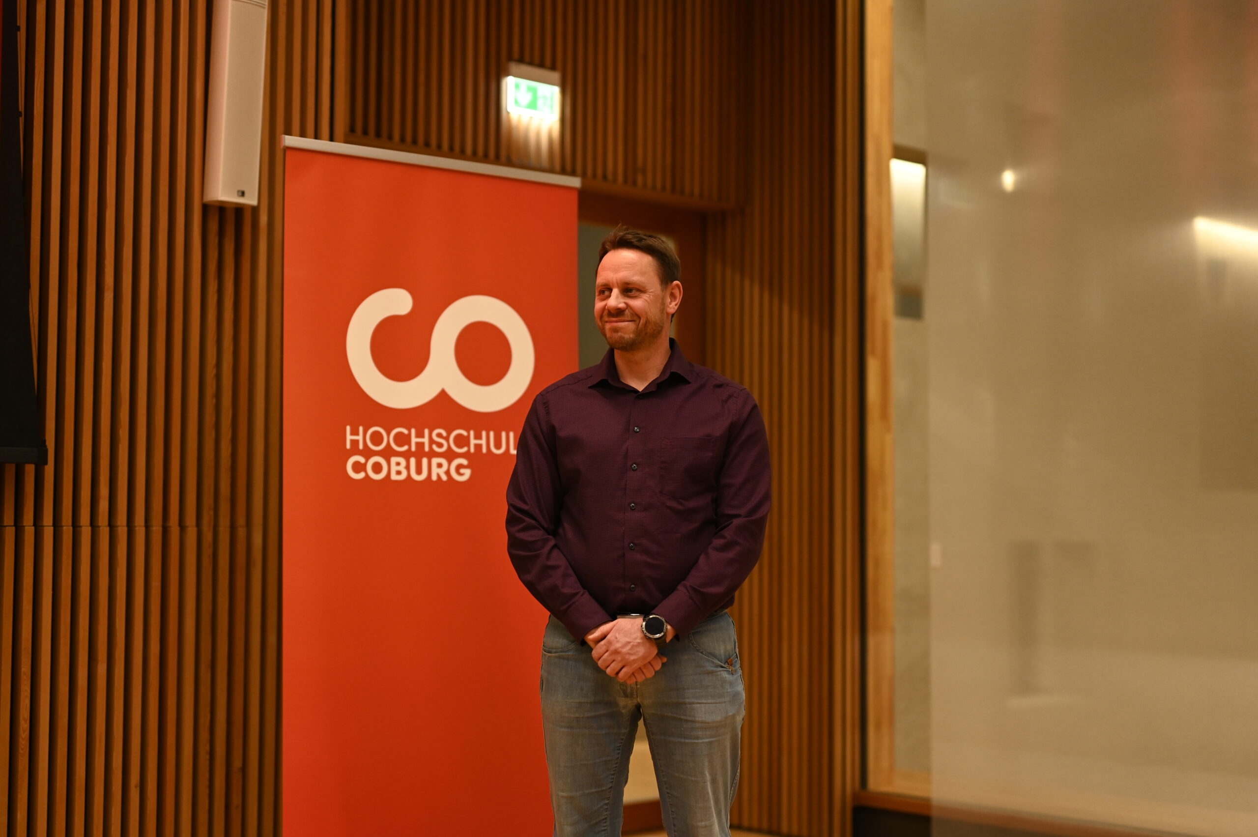 Ein Mann im lila Hemd steht selbstbewusst vor dem großen roten Banner mit der Aufschrift „Hochschule Coburg“, vor dem Hintergrund glatter Holzwände, die das moderne Design des Raumes unterstreichen.