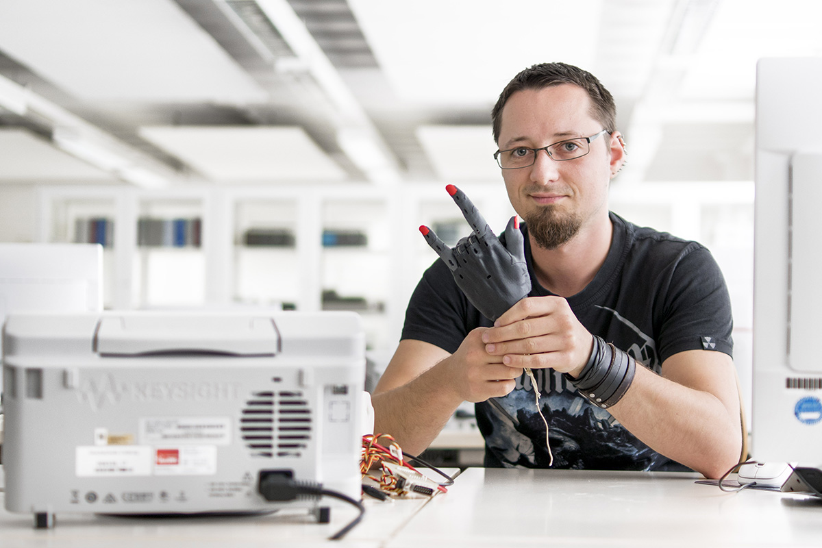 Inmitten der hellen, modernen Arbeitsräume der Hochschule Coburg sitzt eine Person mit Brille und Bart an einem Tisch in einem Labor und hält eine Handprothese in der Hand. Umgeben von elektronischen Geräten verkörpert sie Innovation und Fachwissen in dieser hochmodernen Umgebung.