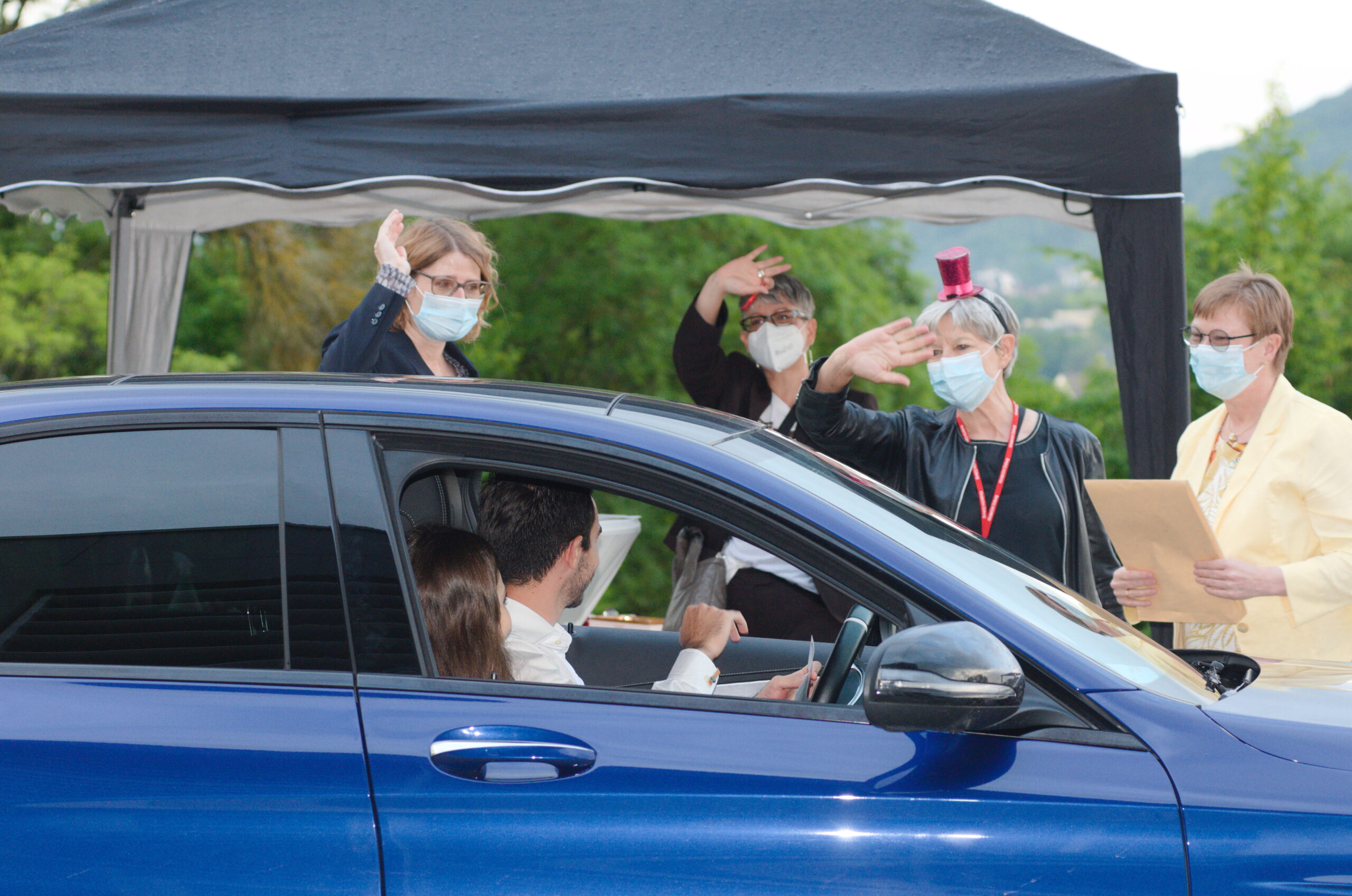 Ein Mann und eine Frau in einem Auto grüßen drei maskierte Frauen, die neben einem Zelt in der Nähe der Hochschule Coburg stehen. Eine Frau mit einem kleinen roten Hut winkt und hält Papierkram in der Hand. Die Szene scheint eine Drive-Through-Veranstaltung mit Bäumen im Hintergrund zu sein.