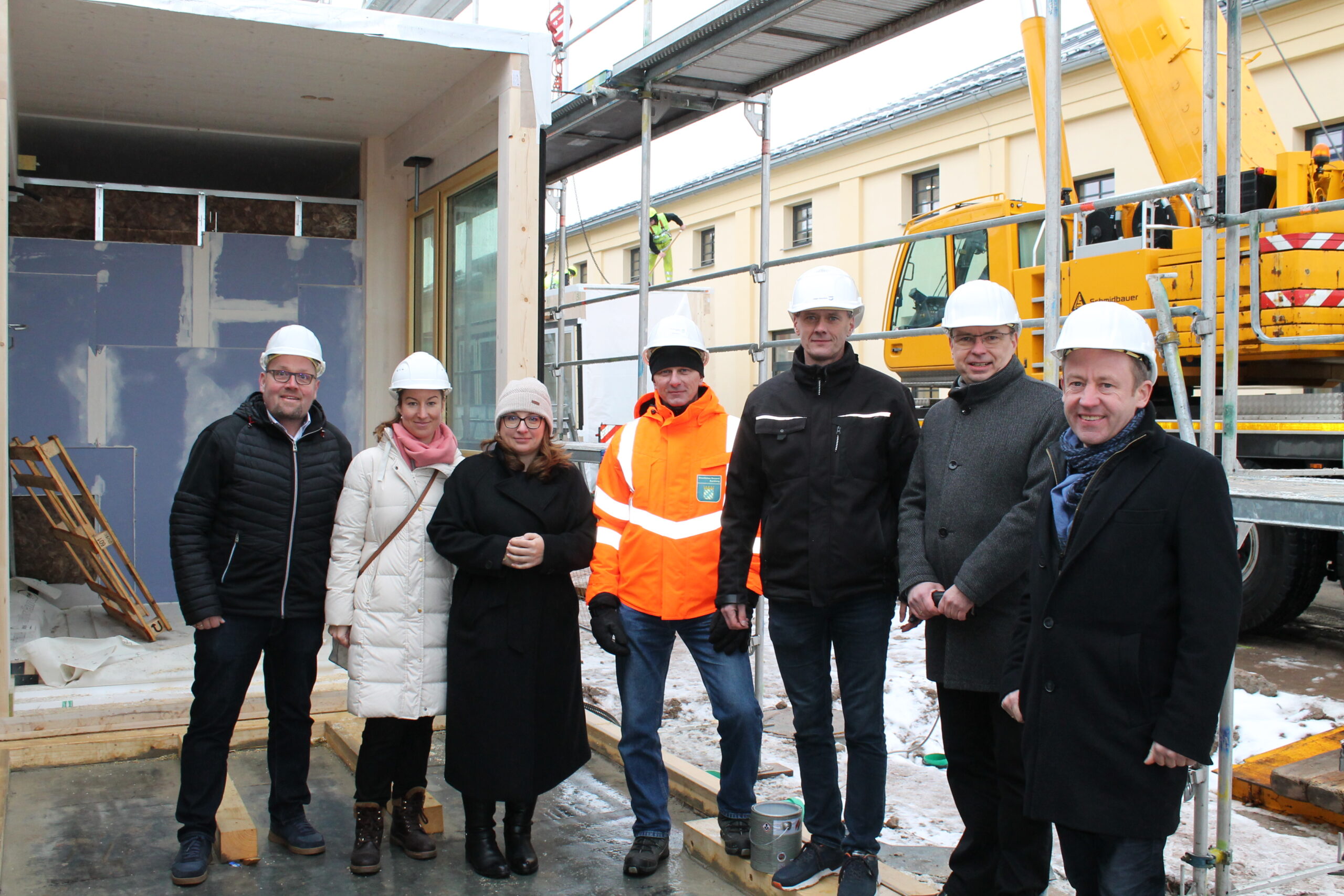 Eine Gruppe von sieben Personen mit Bauhelmen und Winterkleidung steht auf einer Baustelle der Hochschule Coburg. Sie lächeln und scheinen für das Foto zu posieren, im Hintergrund sind Baumaschinen und Gerüste zu sehen.