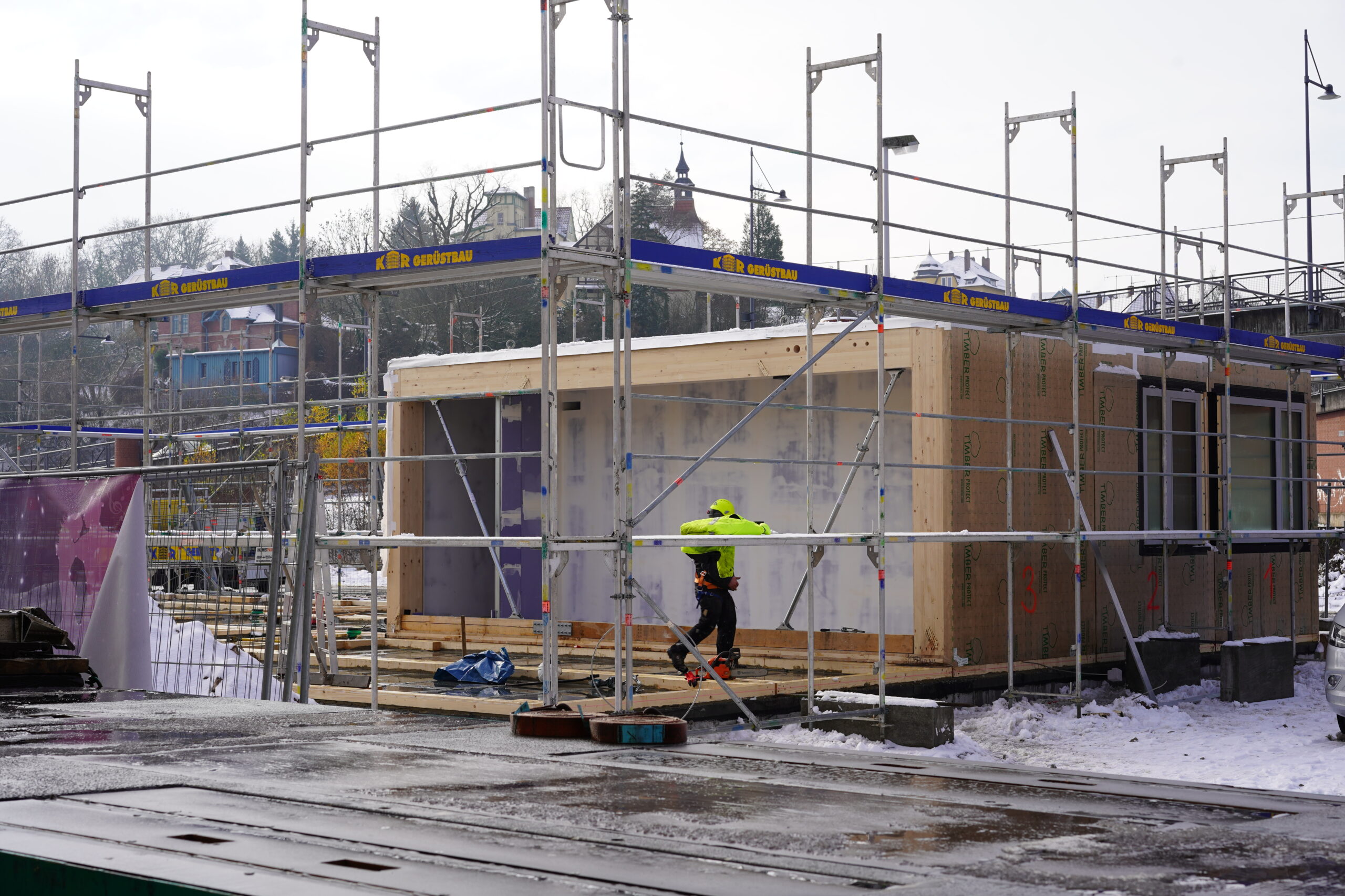 Auf einer Baustelle in der Nähe der Hochschule Coburg ist eine kleine Holzkonstruktion zu sehen, die von einem Gerüst umschlossen ist. Eine Person in einer neongrünen Jacke arbeitet fleißig daneben, während der Boden mit Schnee bedeckt ist und im Hintergrund anmutig Gebäude und Bäume stehen.