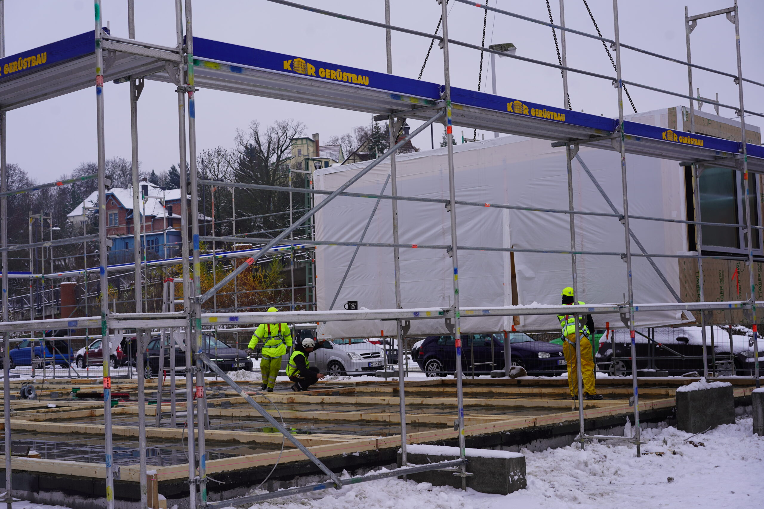 Auf einer Baustelle in einer verschneiten Gegend nahe der Hochschule Coburg sind Arbeiter in gelber und grüner Schutzausrüstung damit beschäftigt, ein Gerüst aufzubauen. Ein großes, weiß bedecktes Objekt dominiert den Hintergrund, dahinter sind Bäume und Wohnhäuser zu erkennen.