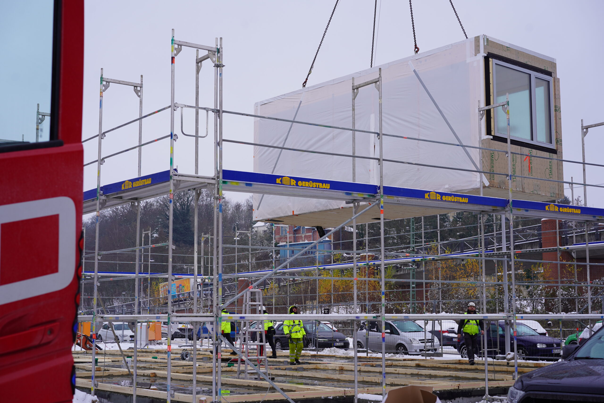 Auf einer Baustelle in der Nähe der Hochschule Coburg wird ein großes vorgefertigtes Gebäudemodul vorsichtig von einem Kran auf ein Gerüst gehoben. Arbeiter in Schutzhelmen und -jacken leiten den Vorgang geschickt, während der Boden und die Umgebung mit Schnee bedeckt sind.