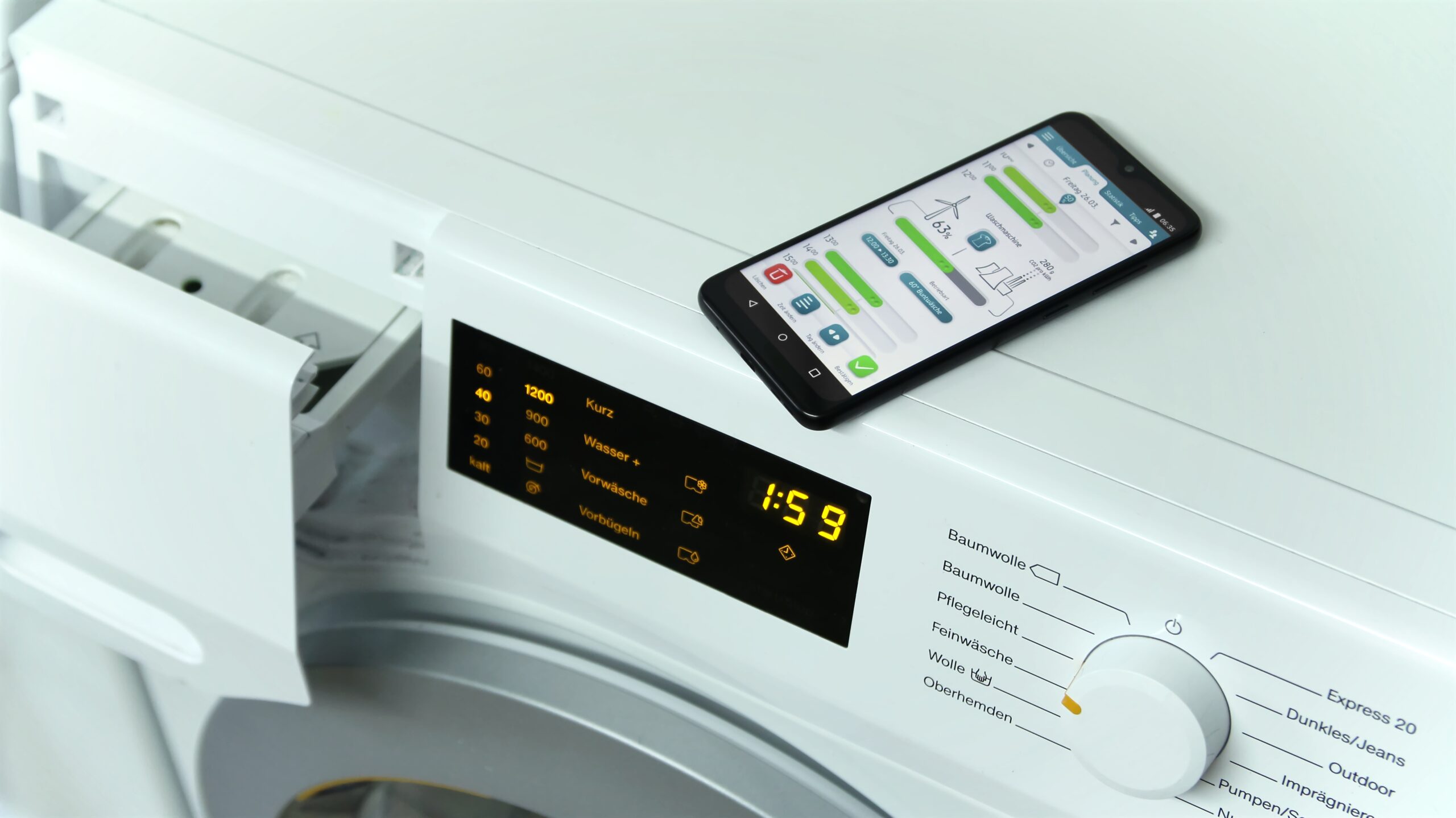 Auf einer Waschmaschine liegt ein Smartphone mit einer Wäsche-App der Hochschule Coburg und zeigt dessen digitalen Bildschirm mit Einstellungen. Links daneben ergänzt eine offene Waschmittelschublade das moderne Design. Die Waschmaschine zeigt eine verbleibende Zeit von 1:59 an.