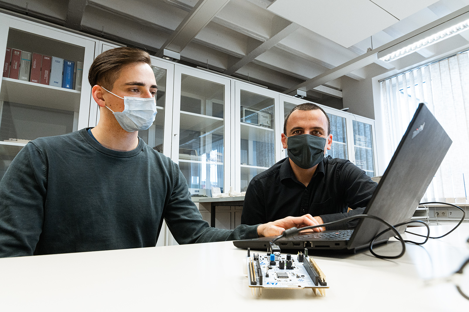 Zwei Männer mit Masken arbeiten in einem Labor der Hochschule Coburg an einem Laptop. Sie konzentrieren sich konzentriert auf ein kleines elektronisches Gerät auf dem Tisch, umgeben von Regalen mit Ordnern und geschlossenen Schränken im Hintergrund.