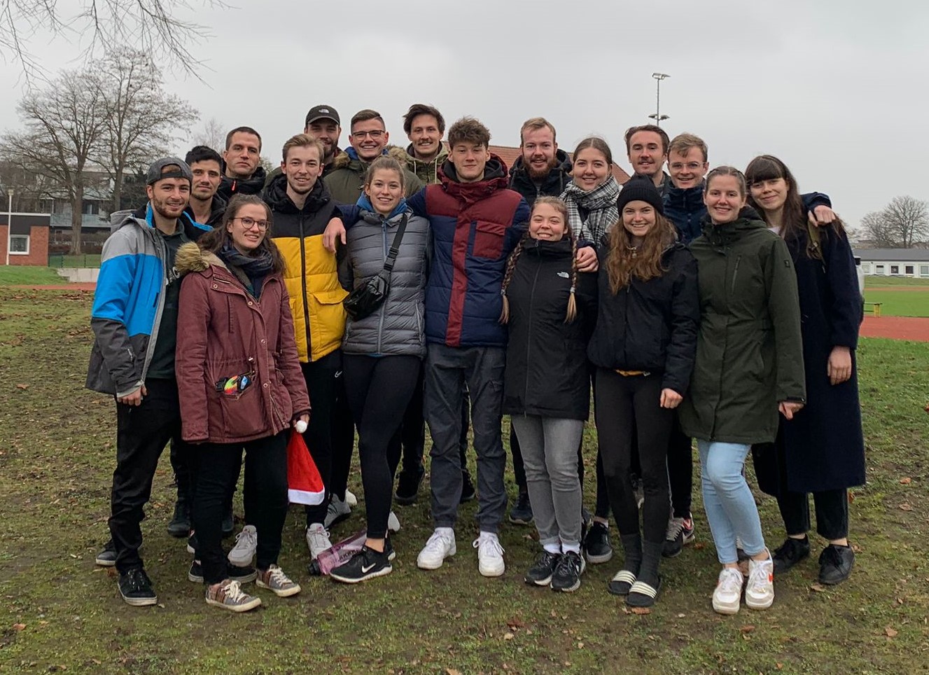 Eine Gruppe von Menschen in Winterkleidung steht gemeinsam auf der Wiese vor der Hochschule Coburg. Sie lächeln in die Kamera, umgeben von Bäumen und einem Sportplatz unter einem bewölkten Himmel.