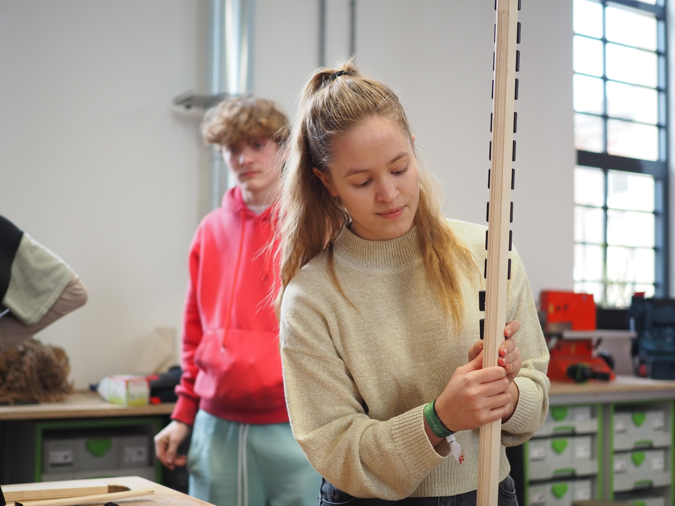 Eine junge Frau in einem beigen Pullover untersucht eine hohe Holzkonstruktion in der Werkstatt der Hochschule Coburg. Hinter ihr beobachtet ein junger Mann in einem roten Kapuzenpullover. Der Raum ist voller Werkzeuge und Materialien auf Regalen und Tischen, alles in natürliches Licht getaucht, das durch die Fenster fällt.