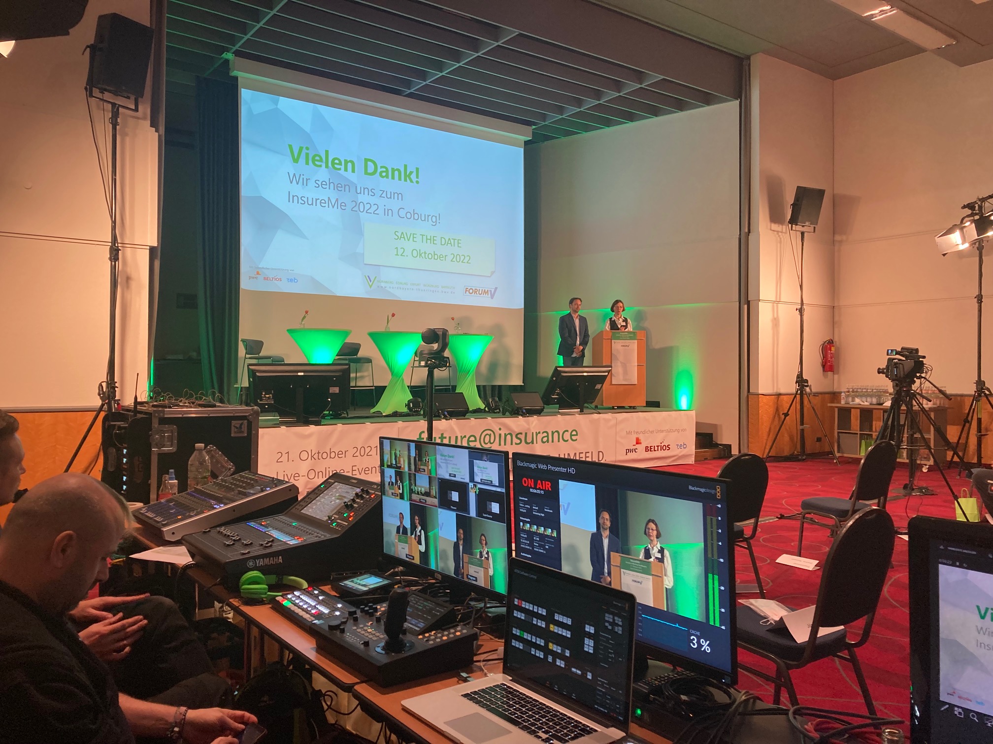 In einem Konferenzraum der Hochschule Coburg steht eine Bühne mit einer Präsentationsleinwand mit der Aufschrift „Vielen Dank!“ Mehrere Personen stehen auf der Bühne, umgeben von hochmoderner Audio- und Videotechnik. Die Atmosphäre ist professionell und technikorientiert.