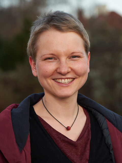 Eine Person mit kurzen Haaren lächelt herzlich, trägt ein kastanienbraunes Hemd und eine dunkle Halskette unter einer offenen Jacke und verkörpert den jugendlichen Geist der Hochschule Coburg. Die verschwommene Naturkulisse im Hintergrund verleiht der Szene eine heitere Note.