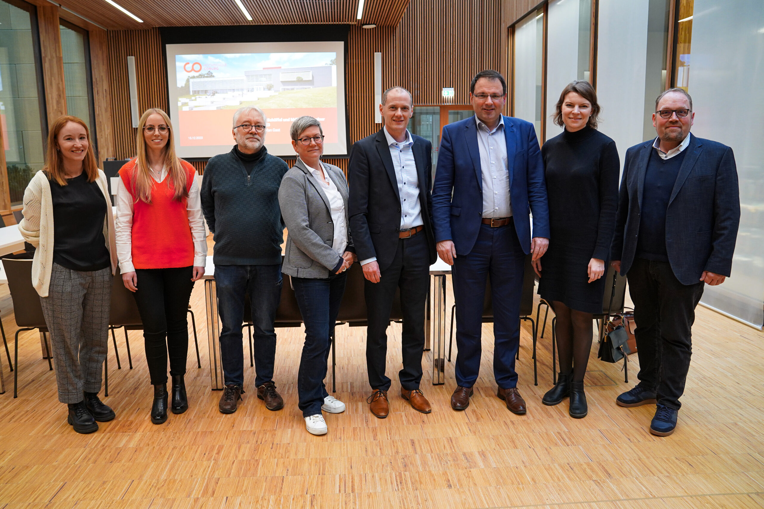 Eine Gruppe von acht Personen steht auf einem Holzboden in einem modernen Raum der Hochschule Coburg. Sie sind in legerer Business-Kleidung gekleidet, im Hintergrund ist ein Präsentationsbildschirm zu sehen.