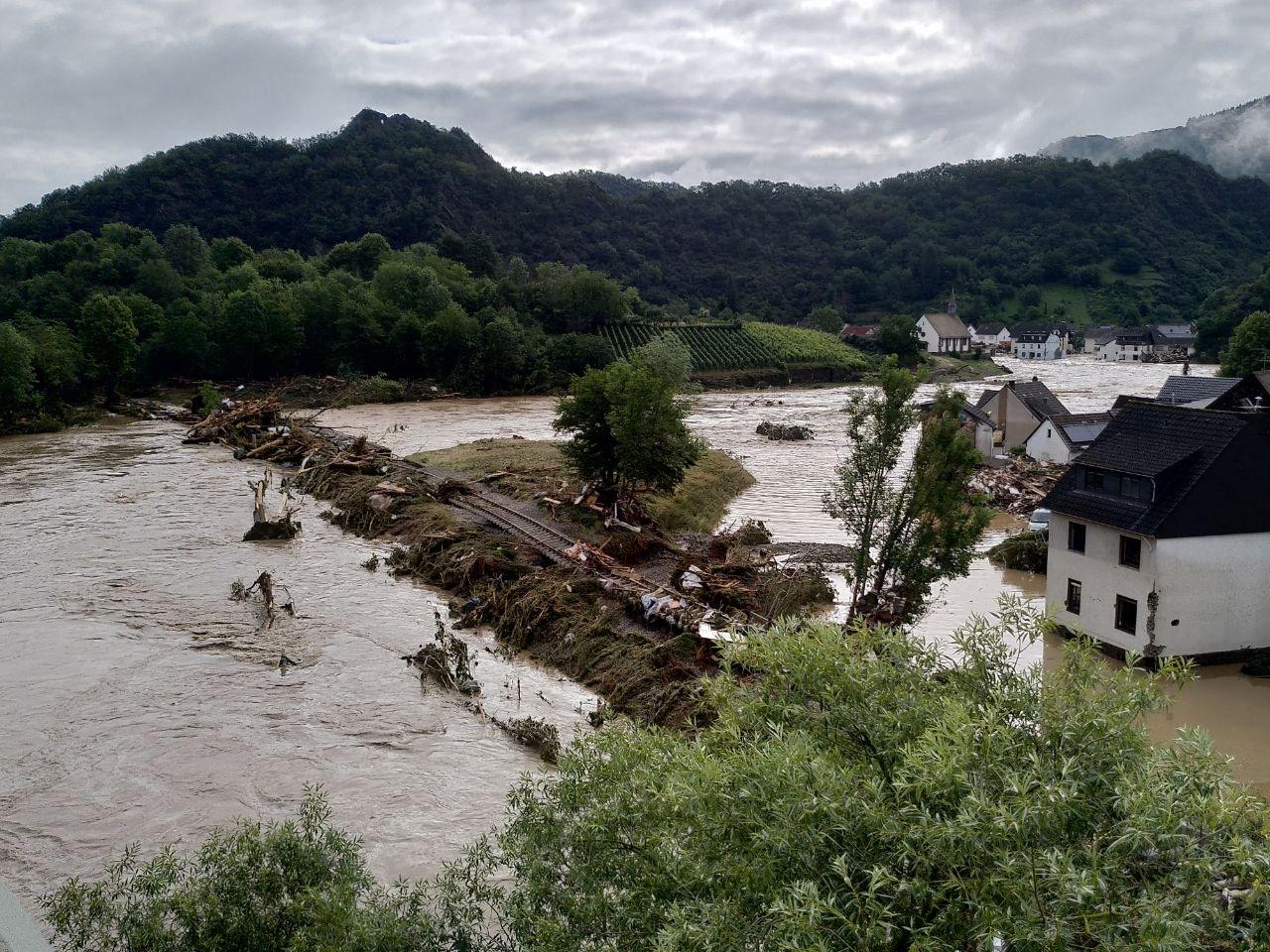 Ein über die Ufer getretener Fluss mit Geröll überflutet die Szene und überschwemmt Gebäude in einem kleinen Dorf in der Nähe der Hochschule Coburg. Saftig grüne Hügel bilden den Hintergrund, während der bewölkte Himmel auf heftige Regenfälle der letzten Zeit hindeutet.