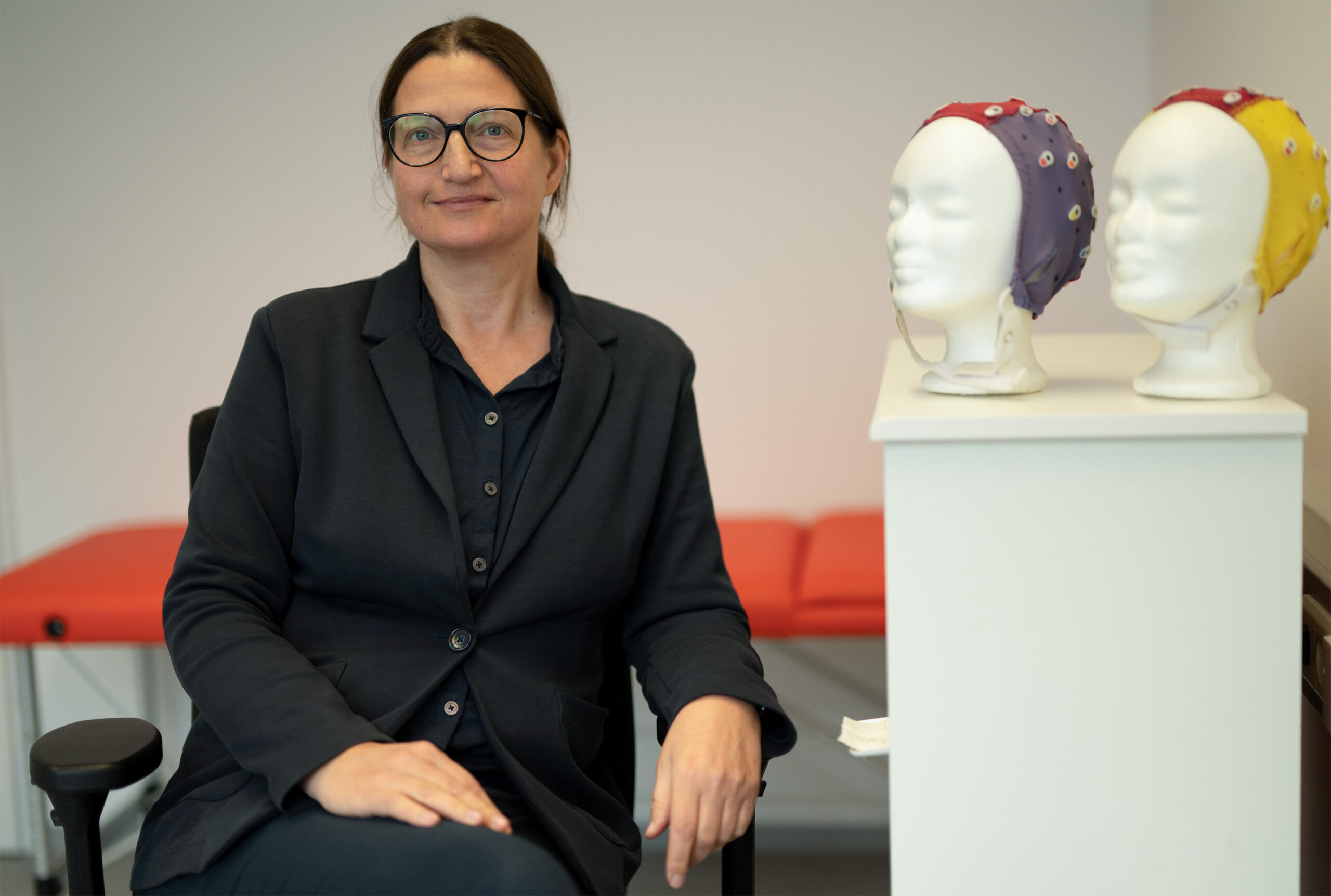 Eine Person im schwarzen Anzug sitzt in einer büroähnlichen Umgebung an der Hochschule Coburg. In der Nähe stehen zwei mit bunten EEG-Kappen geschmückte Puppenköpfe. Im Hintergrund sorgt eine rote Bank für einen Farbtupfer in der Szene.