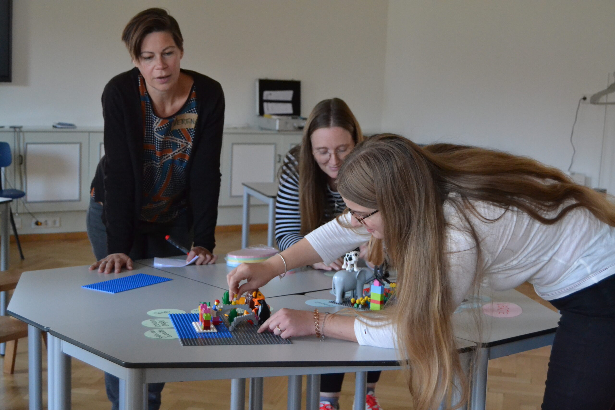 Drei Frauen sitzen an der Hochschule Coburg um einen Tisch mit bunten LEGO-Steinen. Eine Frau beugt sich vor und legt einen Stein hin, während die anderen zuschauen. Die Kulisse scheint ein Klassenzimmer oder eine Werkstatt mit weißen Wänden und Holzboden zu sein.