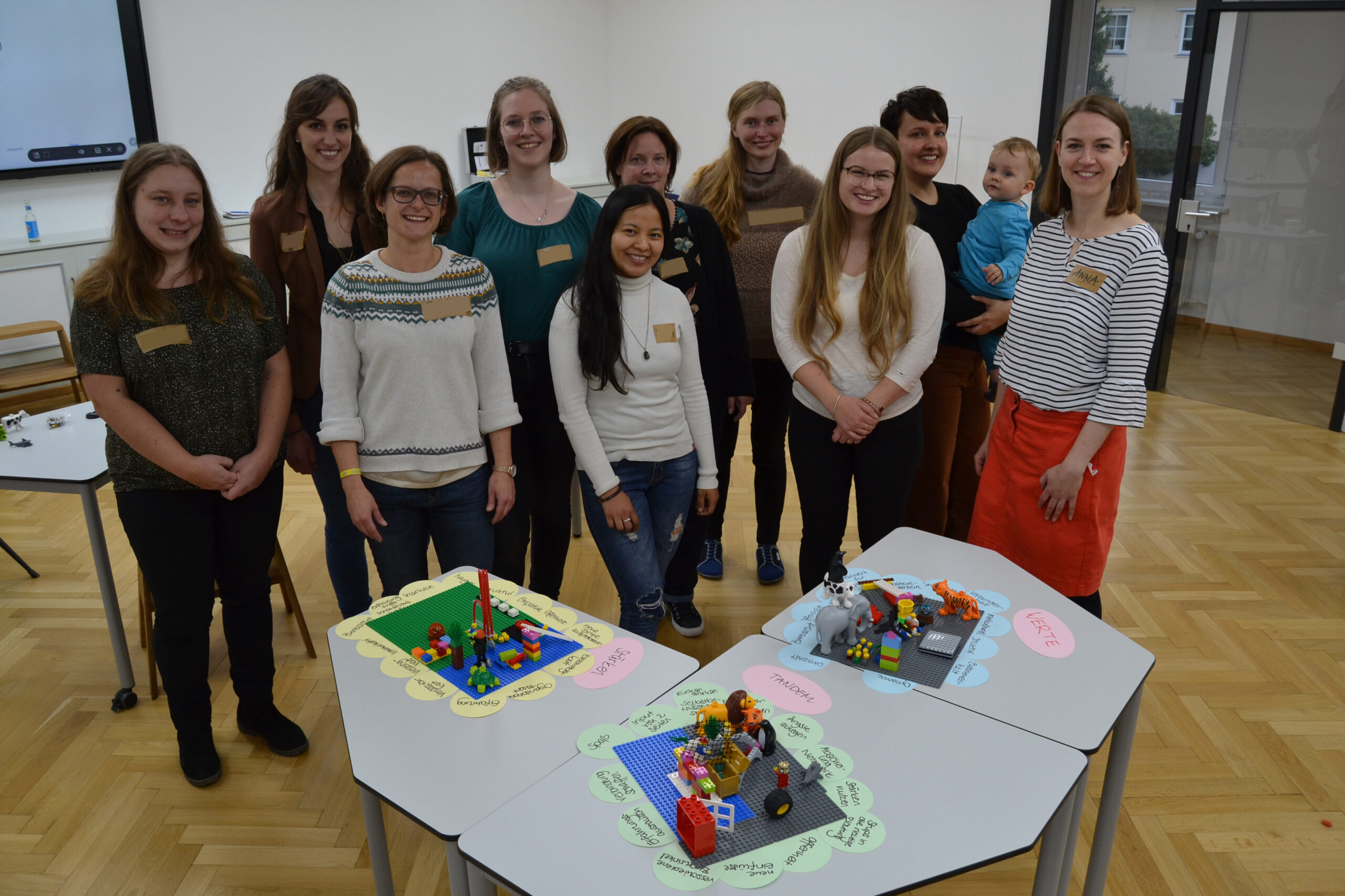 Eine Gruppe von neun Frauen und einem Kind steht lächelnd in einem Raum mit Holzboden in der Hochschule Coburg. Vor ihnen stehen Tische, auf denen bunte LEGO-Modelle auf beschrifteten Papierumrandungen ausgestellt sind. Alle wirken glücklich und leger gekleidet.