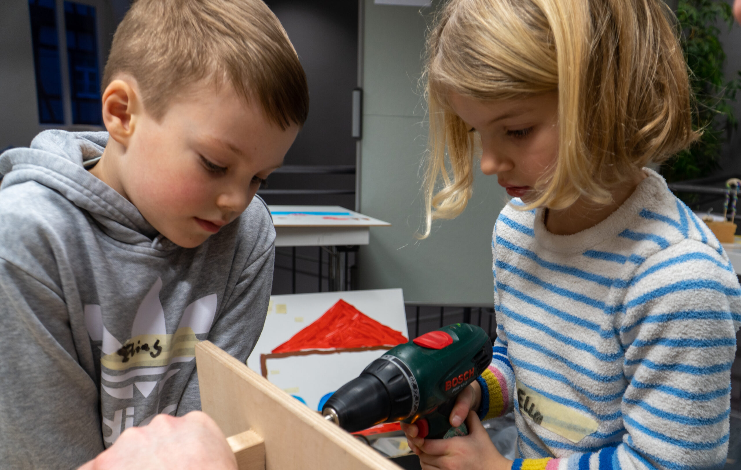 Zwei Kinder, wie angehende Handwerker der Hochschule Coburg, sind in ein Holzbearbeitungsprojekt vertieft. Das Kind links hält ein Holzstück, während das andere eine Bohrmaschine benutzt. Beide sind auf die Aufgabe konzentriert, während eine Zeichnung eines Hauses mit rotem Dach dem Hintergrund ihres Arbeitsbereichs Farbe verleiht.