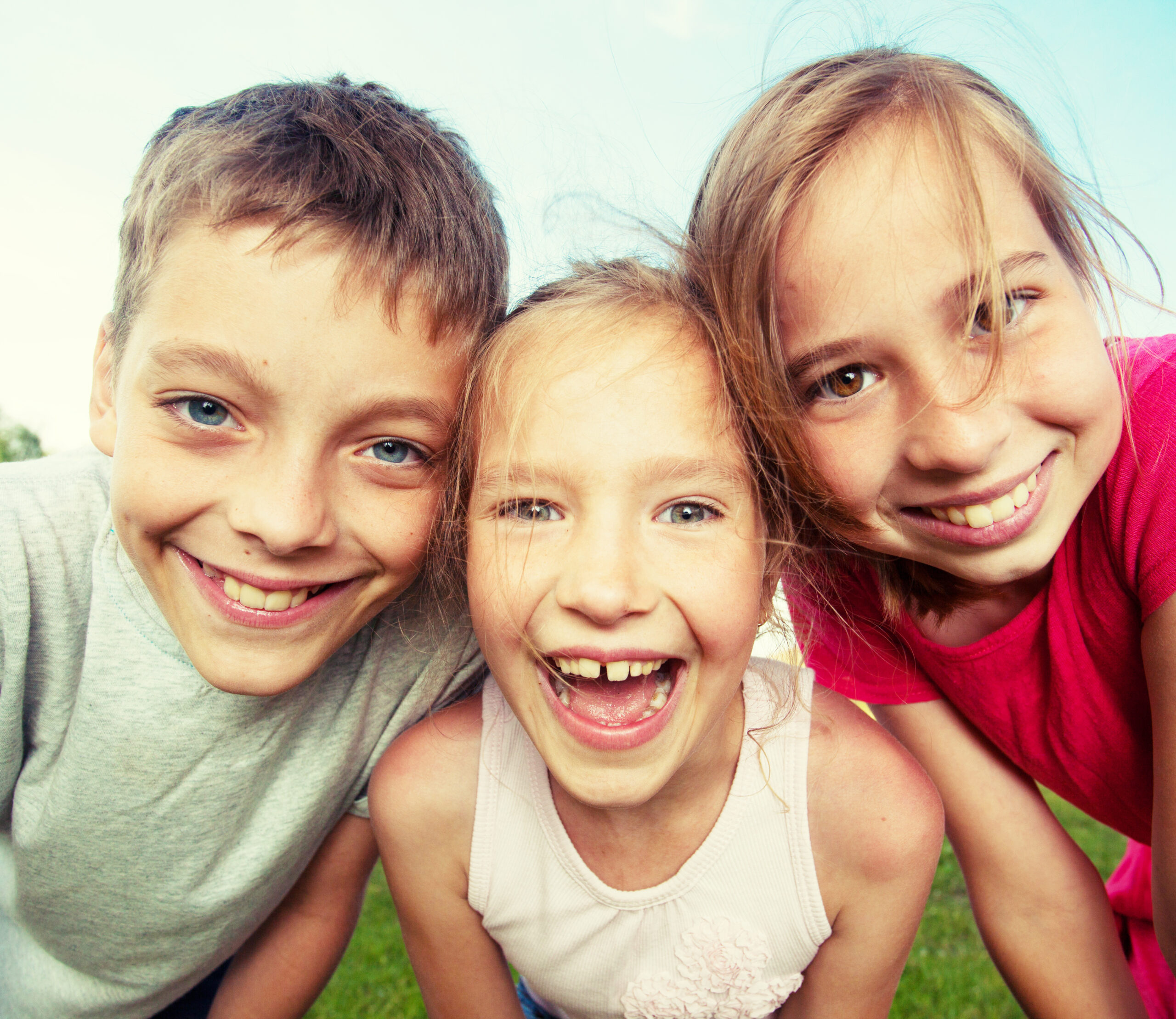 Drei lächelnde Kinder stehen an einem sonnigen Tag im Freien dicht beieinander, mit blauem Himmel im Hintergrund. Sie beugen sich freudig zur Kamera, ihre Freude ist so lebendig und voller Energie wie die eines Studenten der Hochschule Coburg, der sich auf neue Abenteuer einlässt.