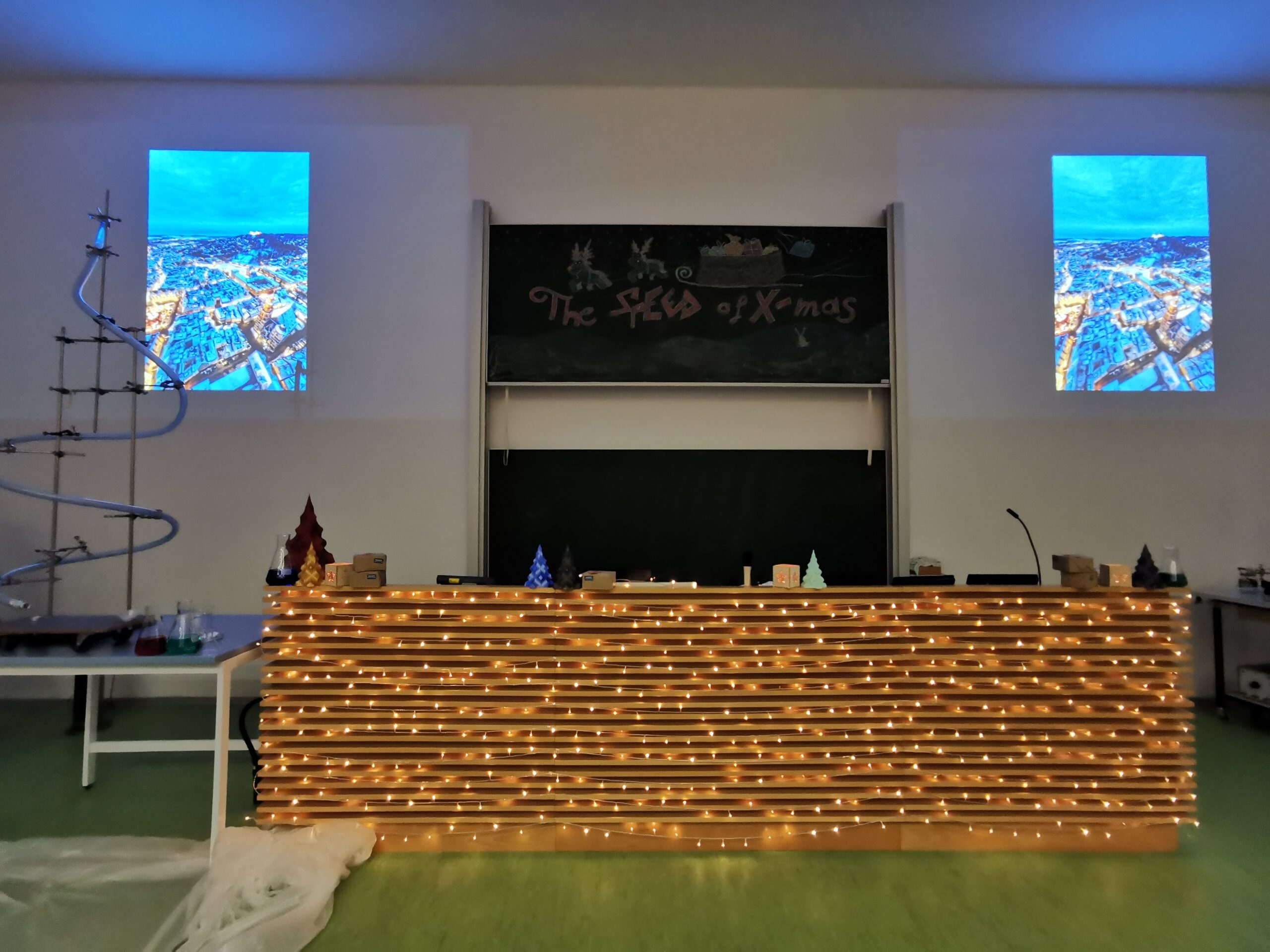 Ein Unterrichtsraum an der Hochschule Coburg besteht aus einem langen Tisch, der mit warmen Lichterketten umhüllt ist. Dahinter steht eine Tafel mit der Aufschrift „The Flow of X-mas“. Darüber zeigen zwei Leinwandprojektionen Stadtansichten aus der Luft, während links ein Achterbahnmodell für spannende Akzente sorgt.