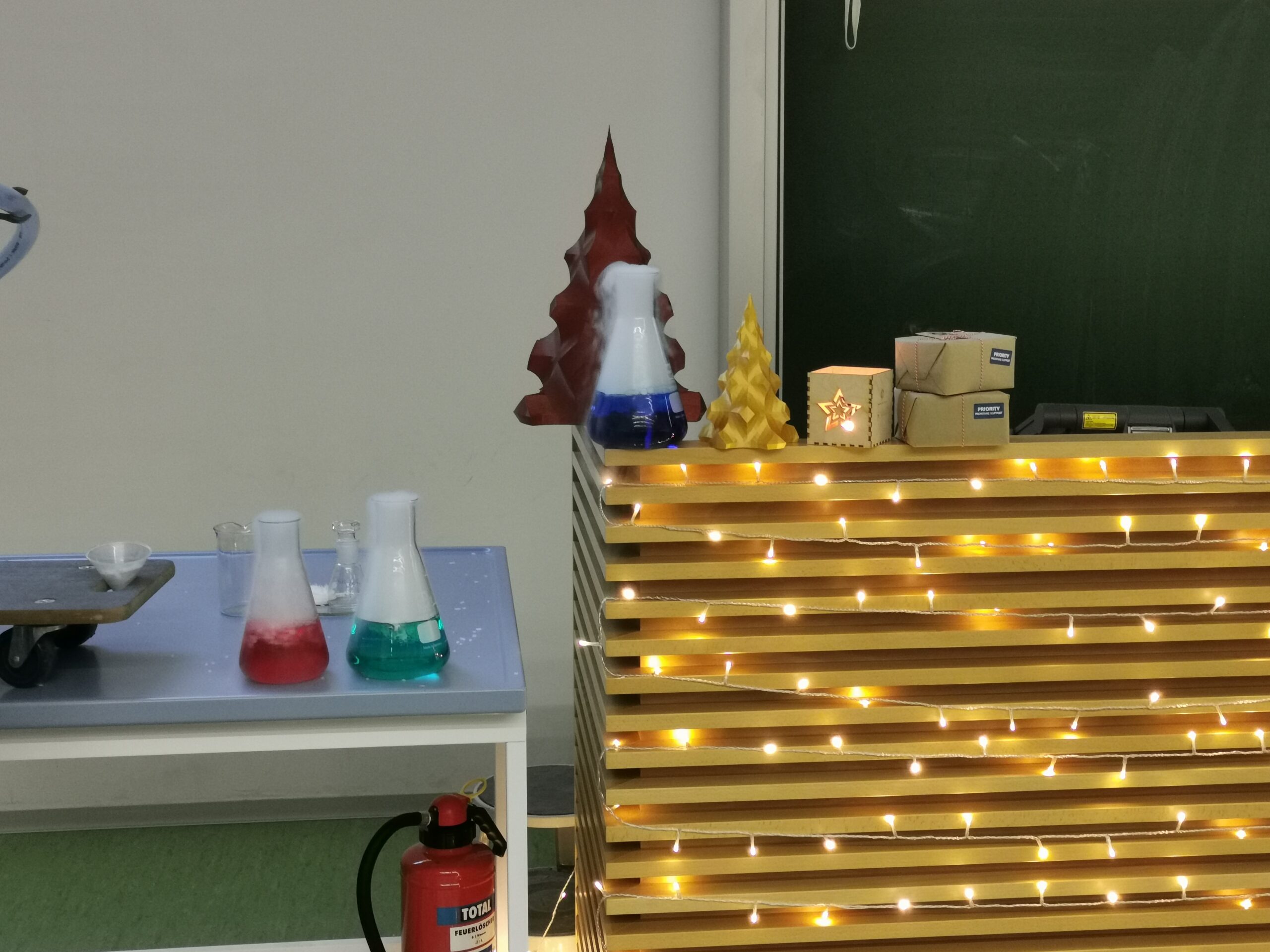 In einem Unterrichtsraum an der Hochschule Coburg steht ein Tisch mit Bechern voller bunter Flüssigkeiten. Lichterketten und Weihnachtsdekorationen schmücken eine Theke, auf der ein roter Weihnachtsbaum, eine gelbe Kerze und eine kleine brennende Laterne zu sehen sind. Auf dem Boden steht ein Feuerlöscher.